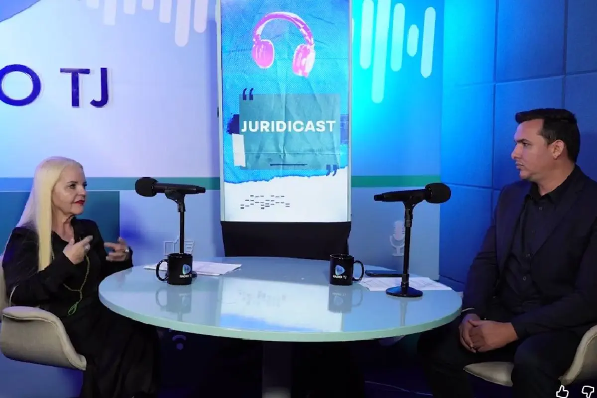 Uma mulher de cabelos longos e claros, vestindo preto, e um homem de terno estão sentados em lados opostos de uma mesa redonda azul em um estúdio de podcast, com microfones e uma tela vertical ao fundo exibindo a logo