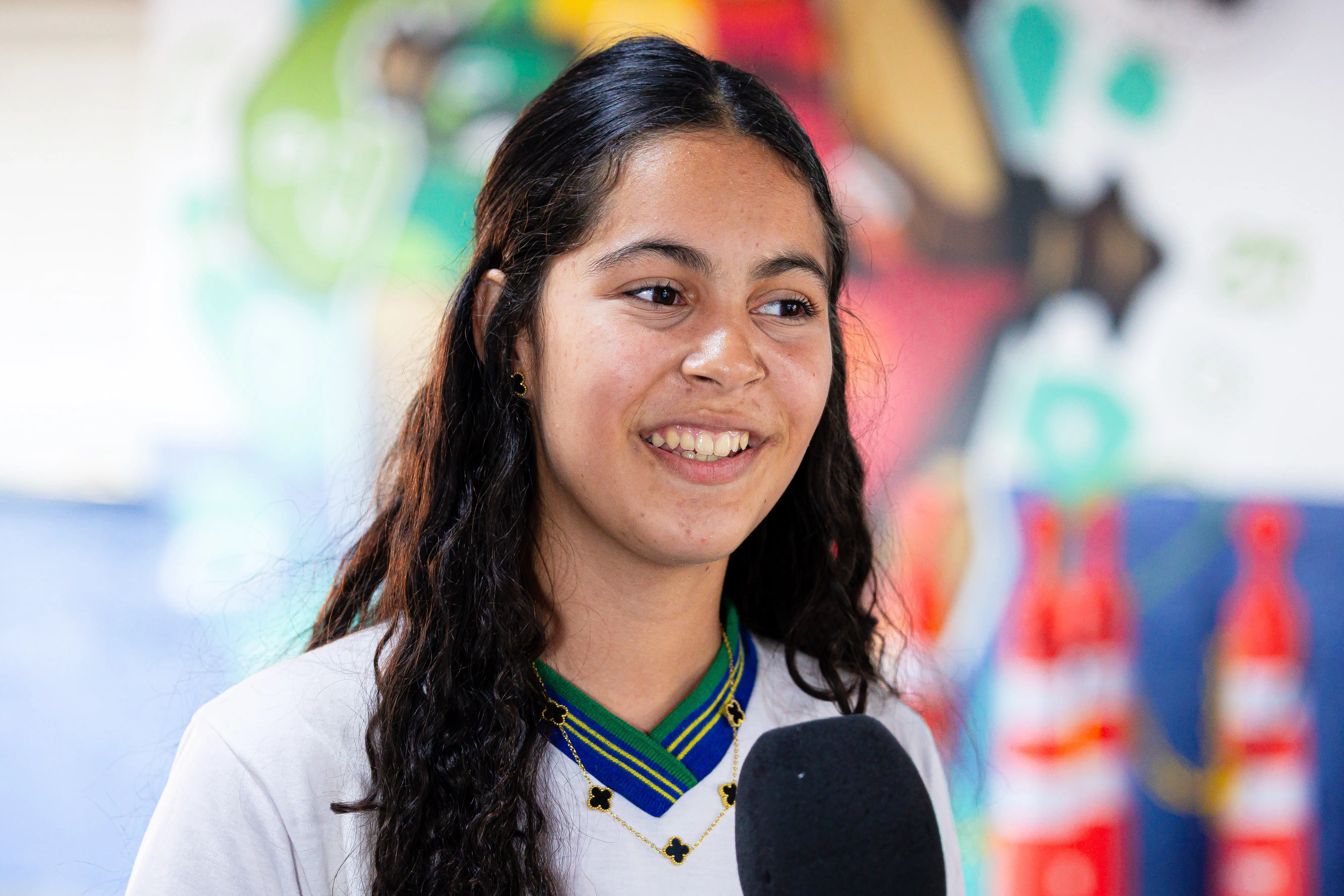 Estudante de cabelos longos e escuros usa camiseta branca com gola azul e verde, brincos e colar dourados, sorri ao falar ao microfone. Ao fundo, mural colorido desfocado. 