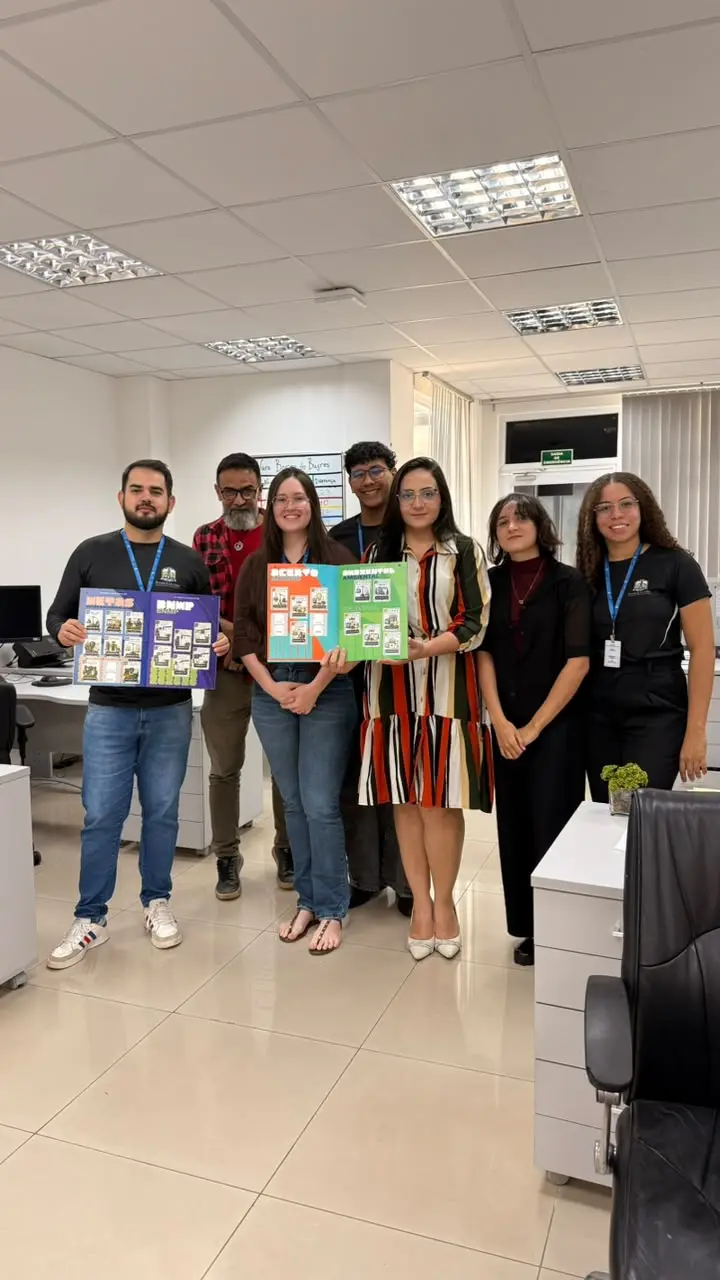 Foto vertical colorida da equipe da 3ª Vara da Comarca de Barra do Bugres posa para foto. O grupo está reunido em pé, na sala onde trabalham, enquanto dois integrantes exibem exemplares abertos do álbum físico da Copa do Judiciário. 