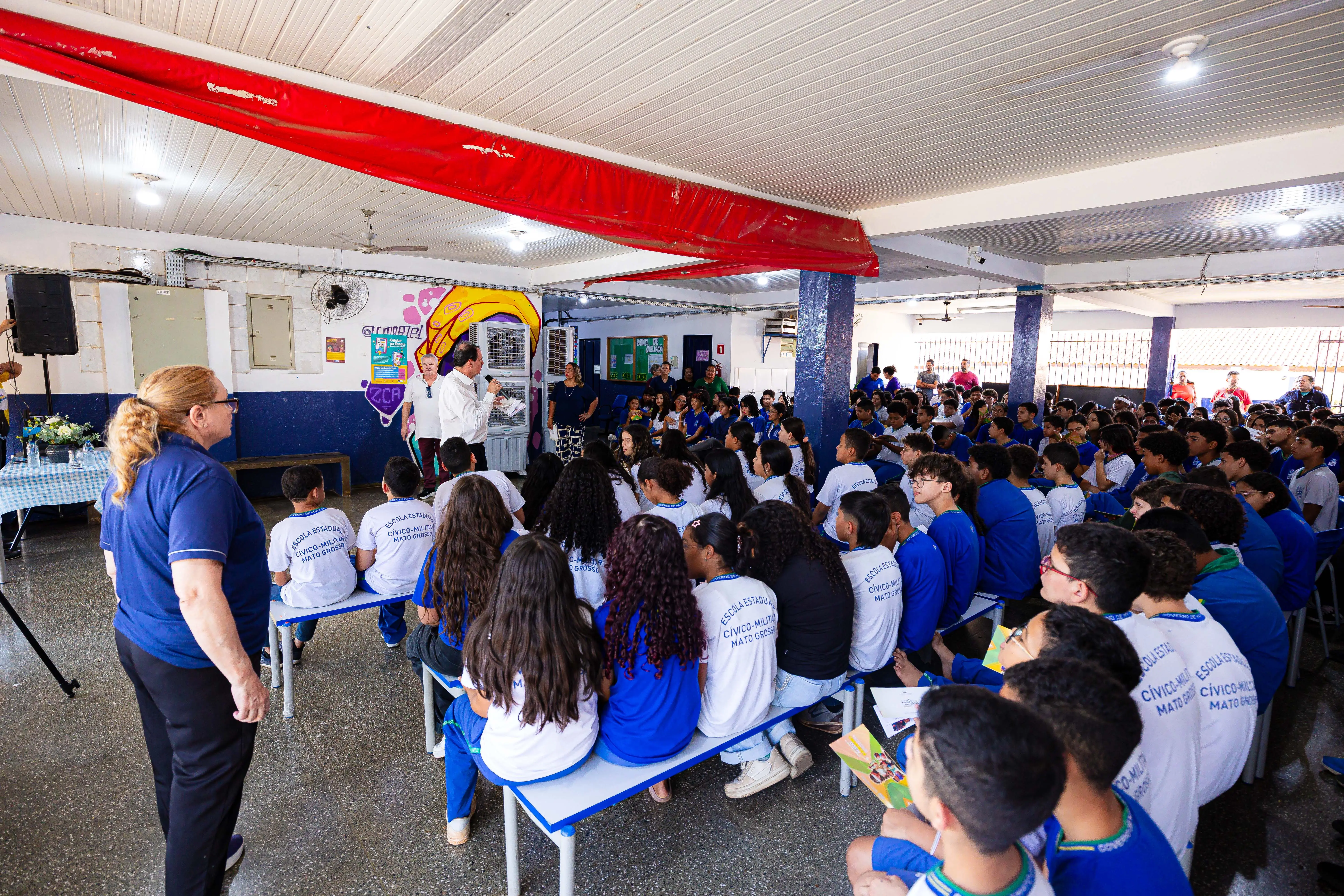 Estudantes uniformizados, sentados em bancos, acompanham atentamente a fala de palestrante durante evento escolar. Ao fundo, faixa vermelha no teto e mural colorido