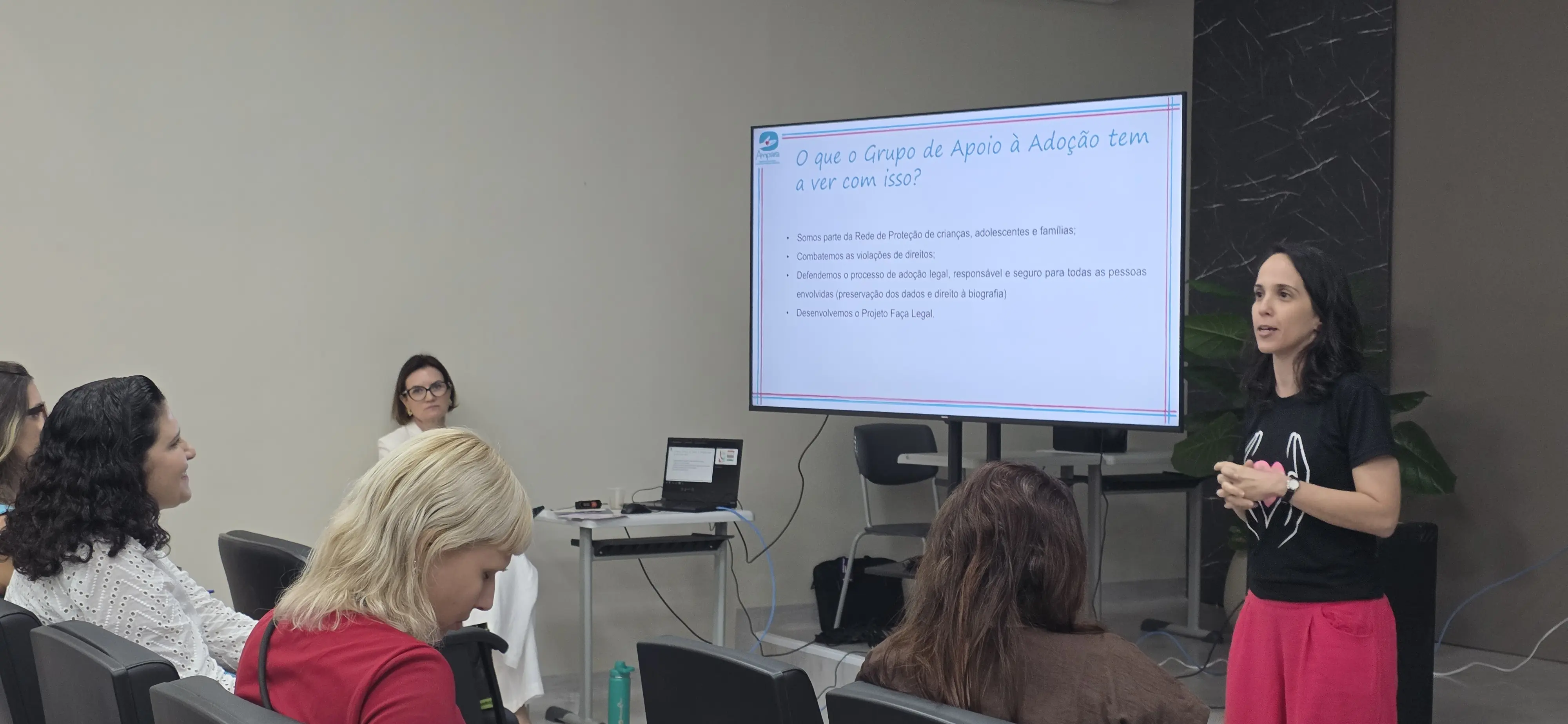 A assistente social da Ampara, Denise Araújo está em pé, à frente do auditório, ela apresenta informações sobre o trabalho dos grupos de apoio à adoção aos profissionais sentados. Ao fundo, a tela exibe conteúdo da apresentação enquanto o público acompanha a palestra.