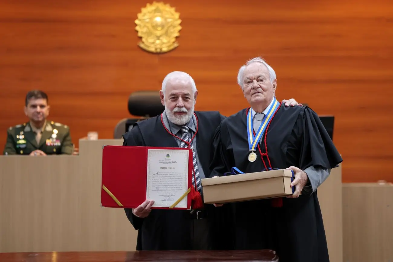 Foto horizontal que mostra o presidente José Zuquim e o desembargador Sérgio Valerio em pé, ao centro do plenário do TJ, abraçados. Zuquim exige um documento em uma pasta vermelha e Valerio segura uma caixa de papel. Eles sorriem para a foto.