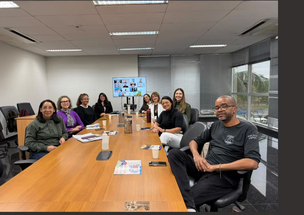 Grupo de dez pessoas sentadas ao redor de uma grande mesa de madeira em uma sala de reuniões. No centro da mesa, um monitor exibe uma videochamada, e o grupo mescla presença física com participantes remotos.