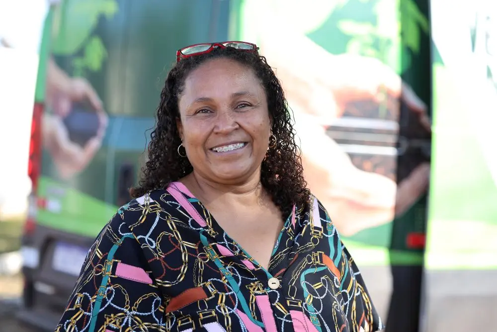 Foto horizontal que mostra a gestora da Escola Municipal Dom Bosco, Odisleia Silva, do busto pra cima, sorrindo para a foto. Ela é uma mulher negra, de cabelos cacheados, castanhos e soltos, usando camisa com estampa colorida e óculos de grau na cabeça. Atrás dela, aparece a van do Verde Novo.