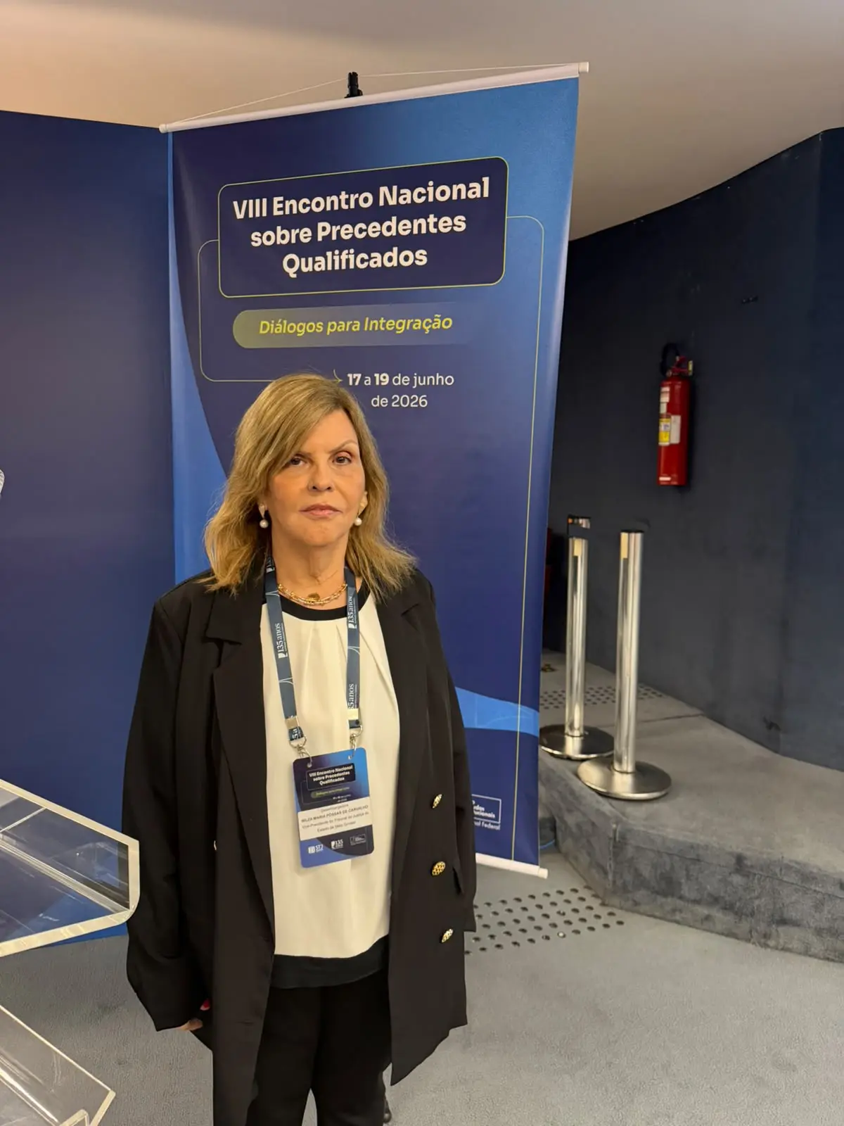 Mulher de cabelos loiros e altura mediana, vestindo blazer preto sobre blusa branca e crachá de identificação, posa em frente a um banner do 