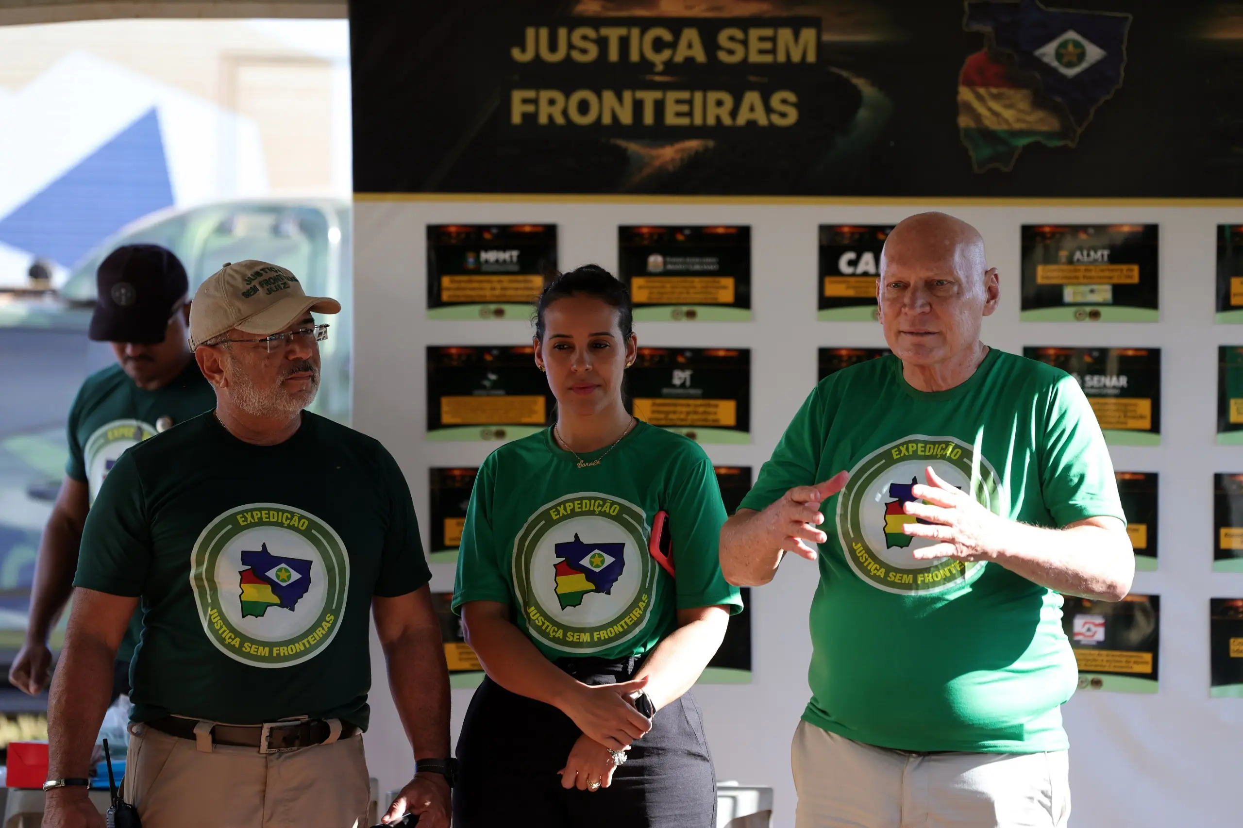 O homem fala gesticulando, ao lado de uma mulher e do homem de boné bege. Todos vestem camisetas verdes da expedição "Justiça Sem Fronteiras". Ao fundo, um painel com informativos.