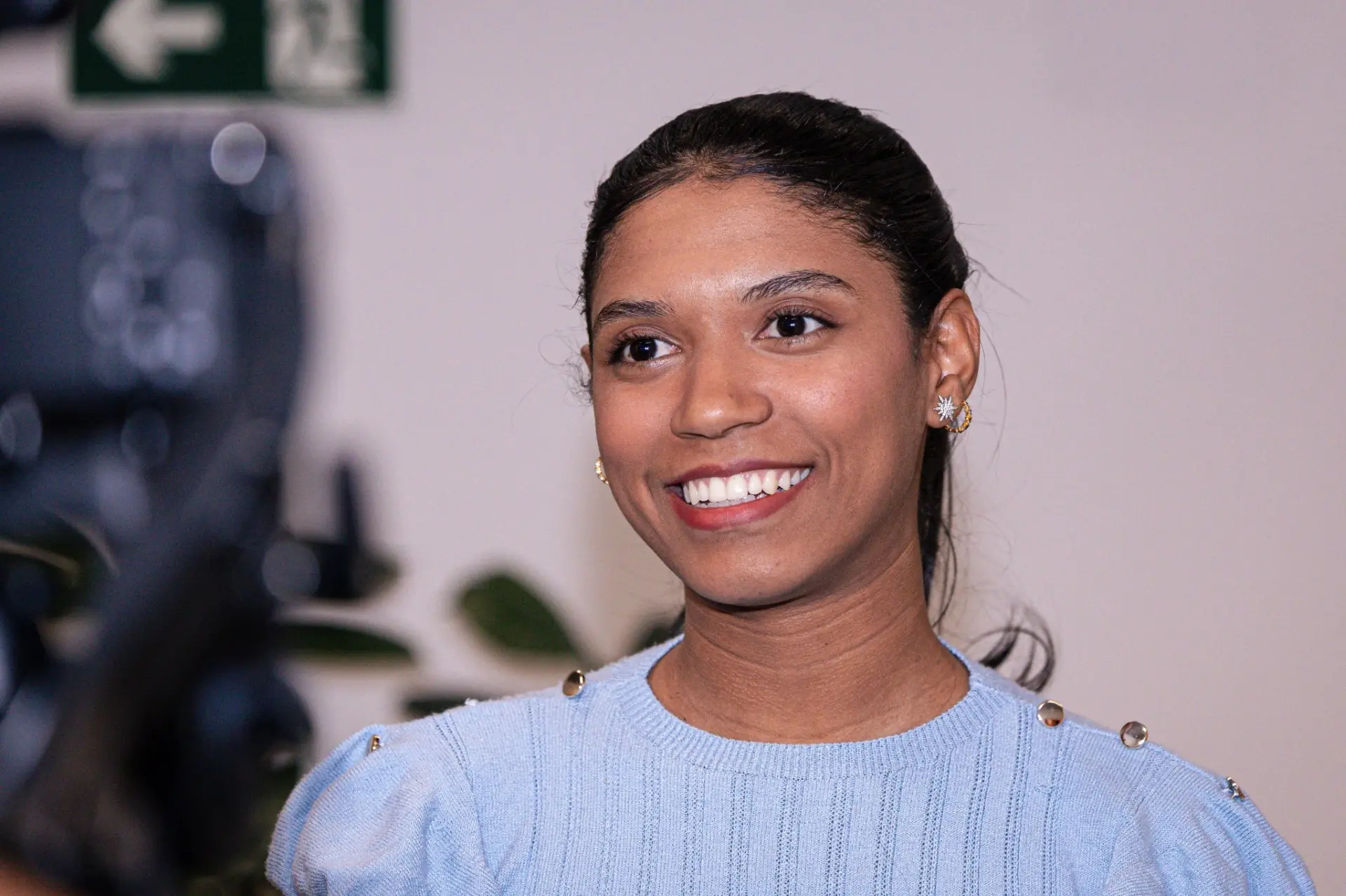 Foto horizontal que mostra a estagiária de Direito Lavínia Darc do busto pra cima, sorrindo, durante entrevista. Ela é uma jovem negra, de cabelos lisos, presos e presos em rabo-de-cavalo, olhos castanhos escuros, usando blusa azul clara e brincos.