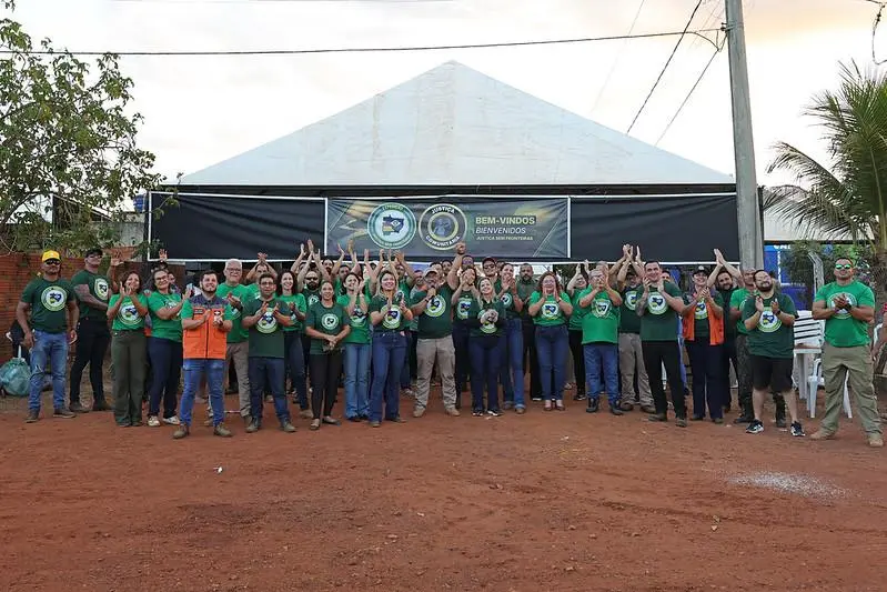 um grande grupo de pessoas com camisetas verdes aplaude em frente a uma tenda branca com uma faixa preta que diz 