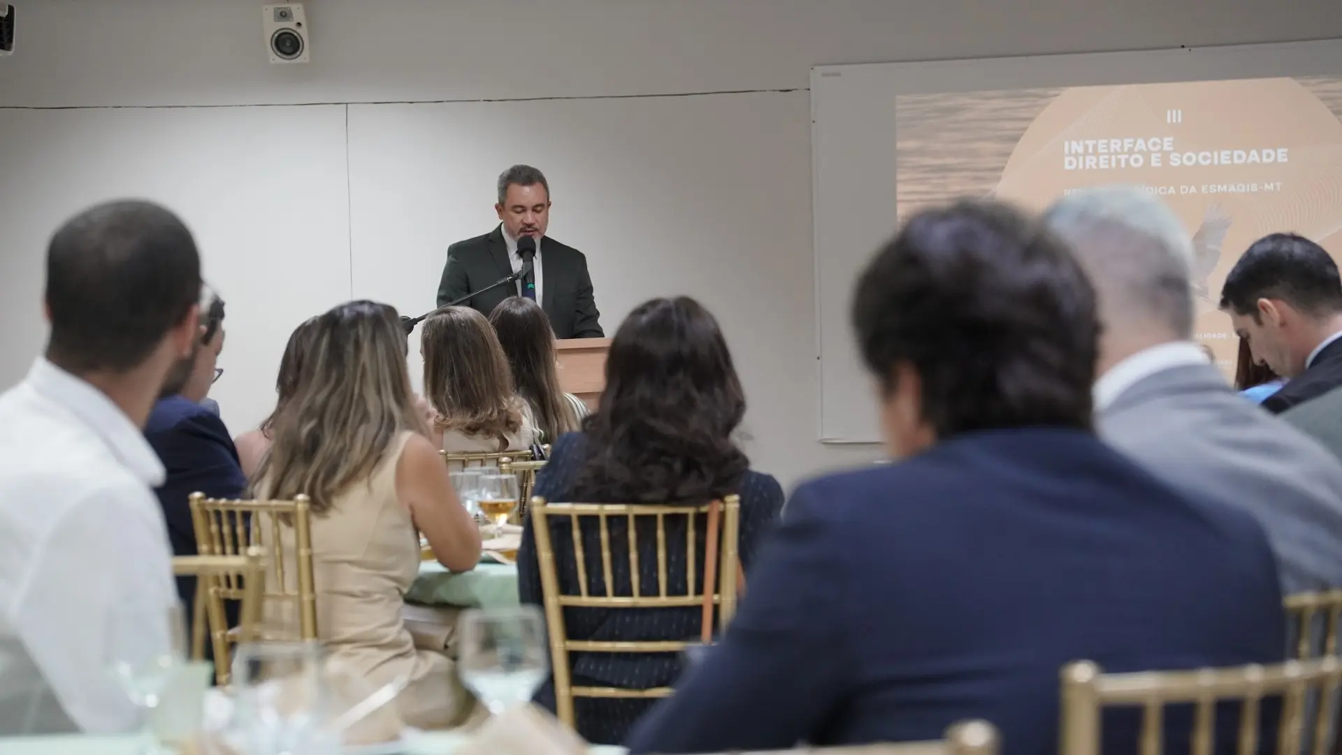 Em ambiente interno, juiz Antonio Peleja, em traje formal, discursa em pé atrás de um púlpito com microfone. Em primeiro plano, aparecem pessoas sentadas em cadeiras, voltadas para a frente. Ao fundo, há parede clara e iluminação uniforme, caracterizando um evento institucional ou acadêmico.