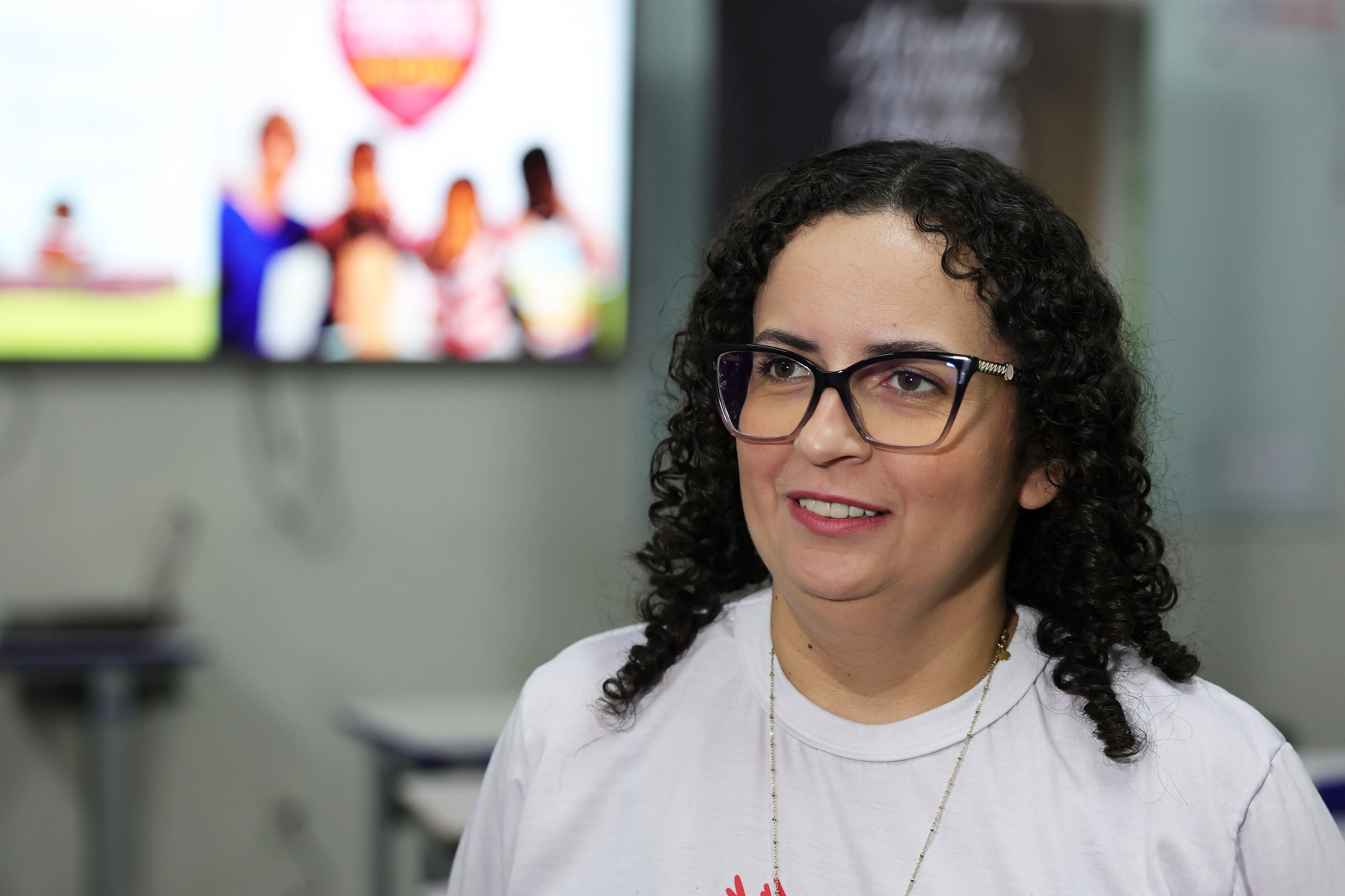 Close-up de mulher de óculos de grau e cabelos cacheados escuros vestindo camiseta branca, olhando para o lado e sorrindo levemente em ambiente interno.