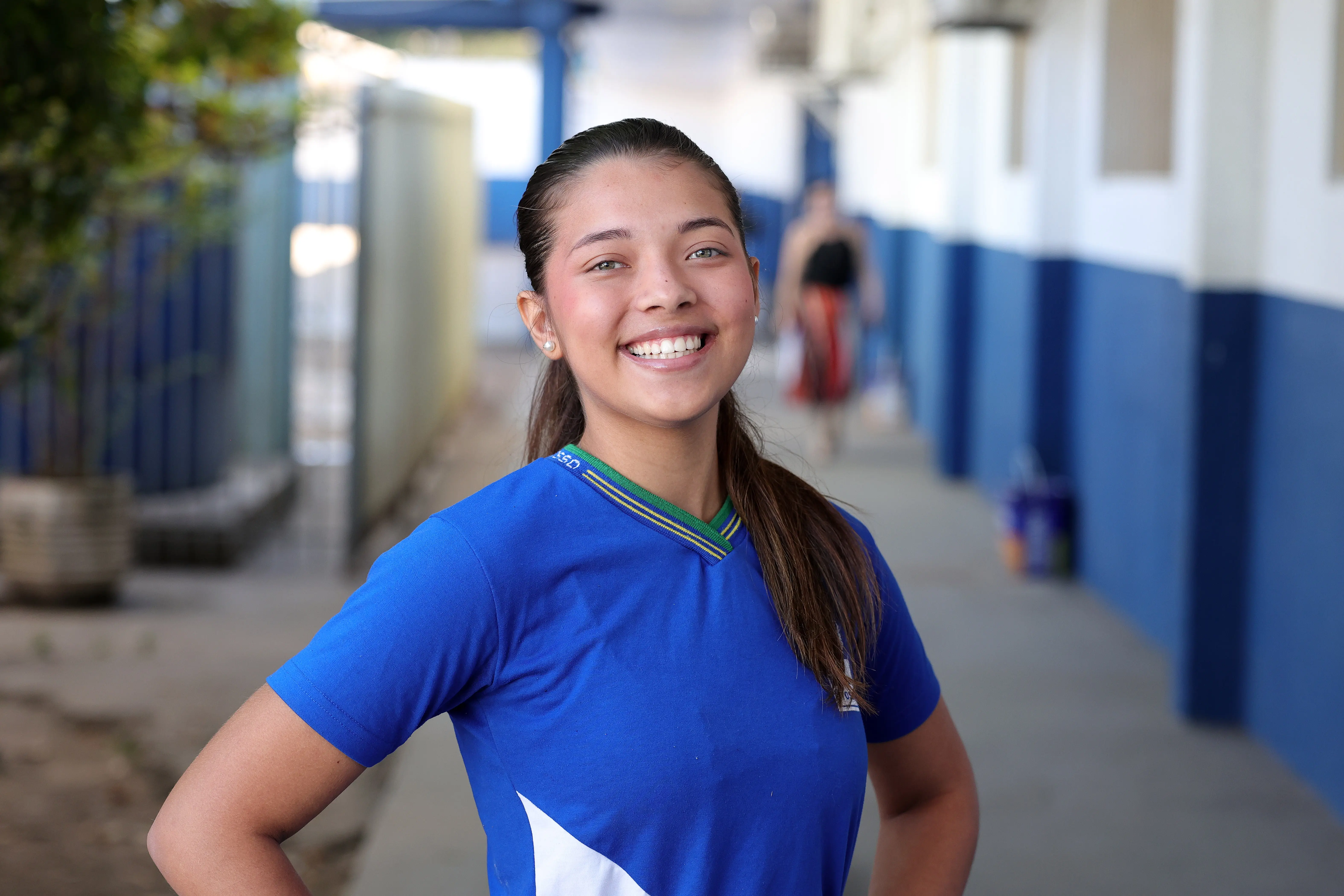 Jovem sorridente com cabelos castanhos amarrados em rabo de cavalo, vestindo camiseta azul de uniforme escolar, posando em um corredor ao ar livre.