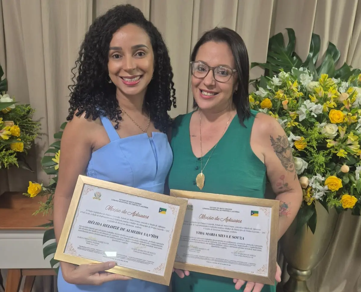 Duas mulheres sorridentes posam lado a lado segurando certificados em molduras douradas (