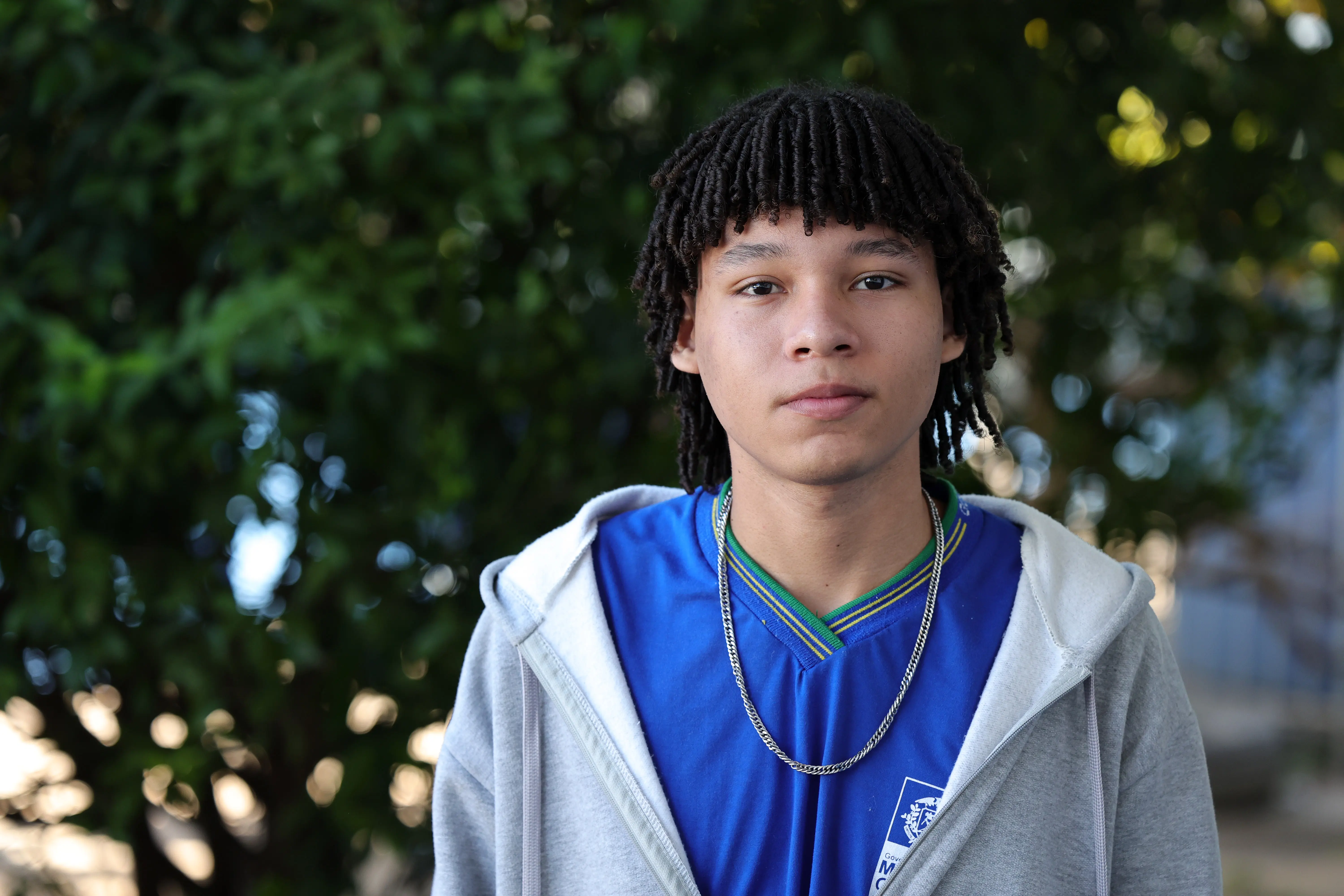 Retrato frontal de um jovem com cabelo cacheado estilo dreads curtos, vestindo jaqueta cinza aberta sobre uniforme escolar azul e uma corrente no pescoço.
