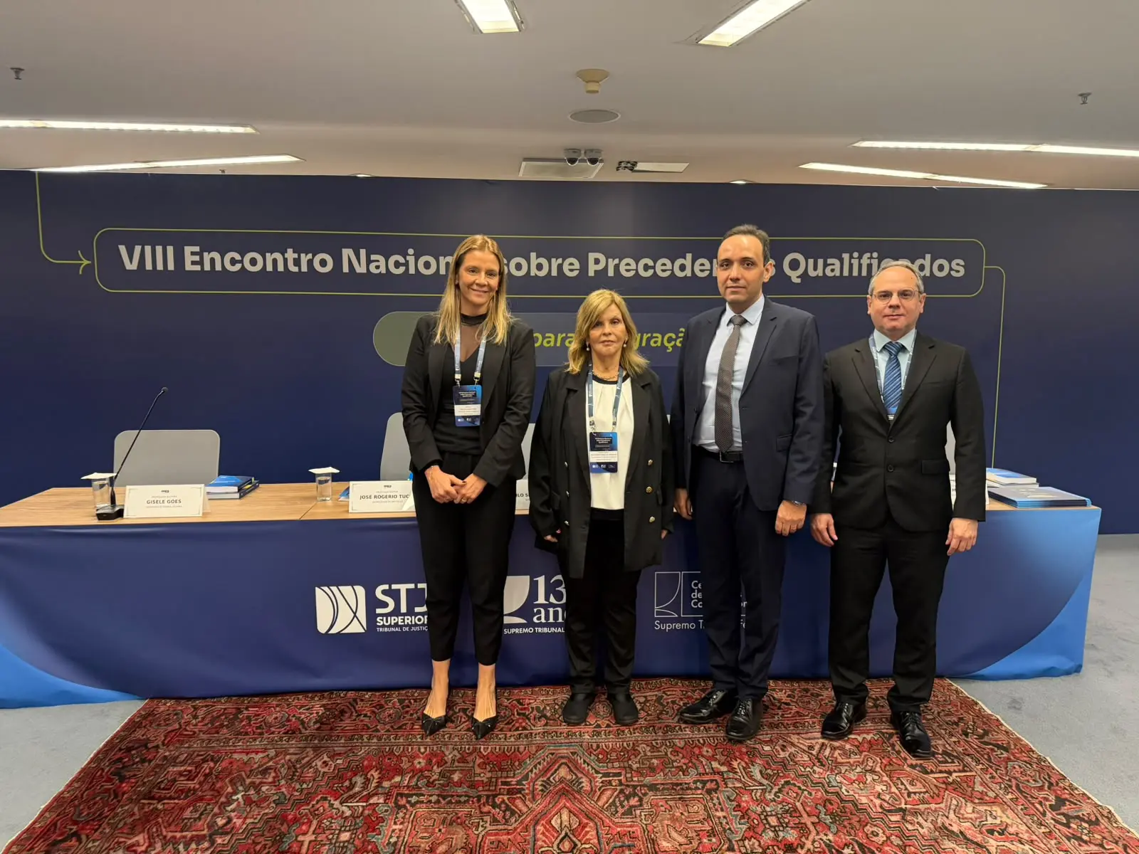 Quatro pessoas em traje social posam lado a lado em um palco. Atrás delas, uma parede azul exibe o título 