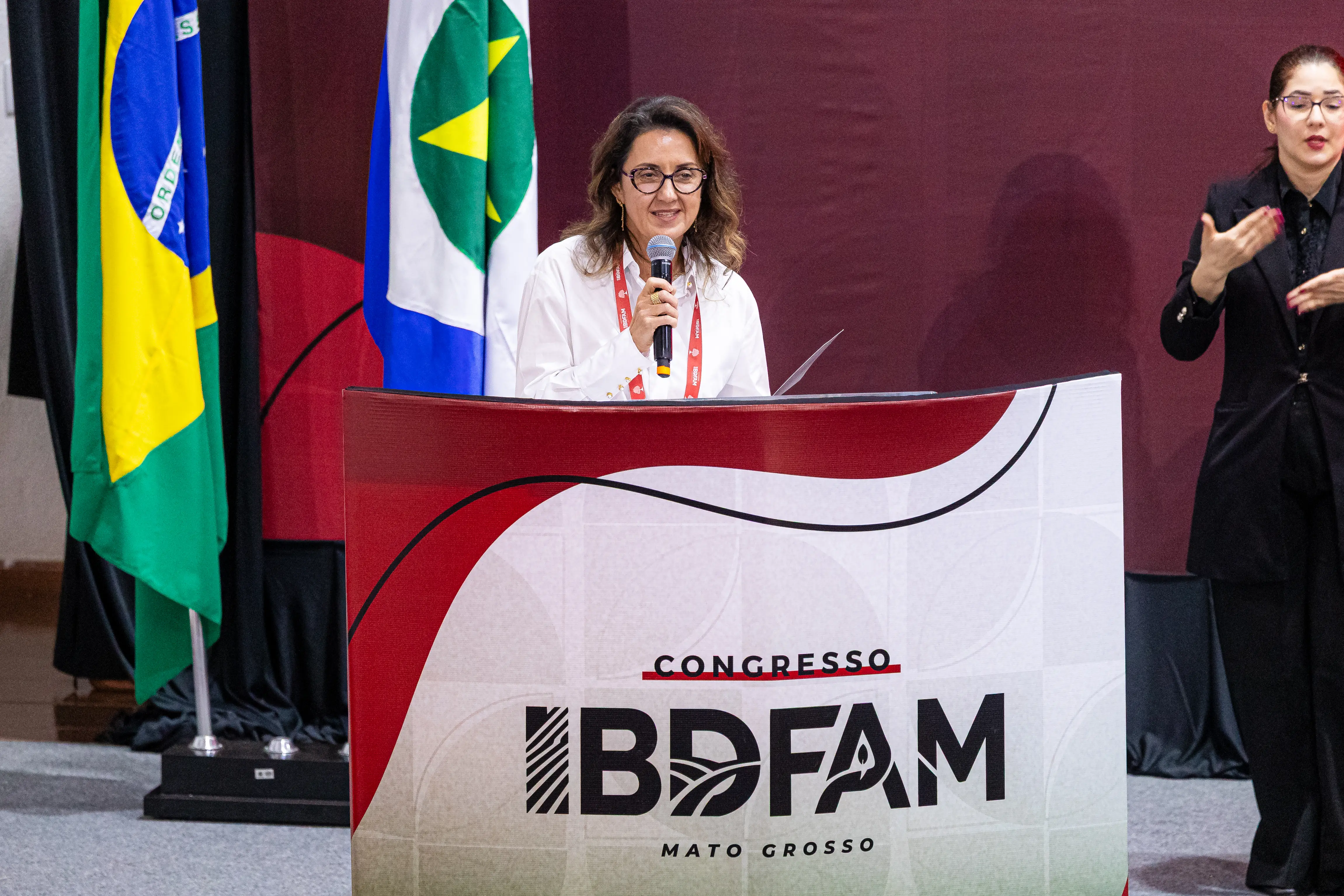 Mulher de óculos e camisa branca fala ao microfone em um púlpito com o logotipo do Congresso IBDFAM Mato Grosso. Ao lado, uma intérprete de Libras e, ao fundo, as bandeiras do Brasil e do estado.