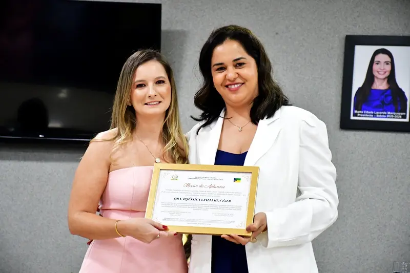 Duas mulheres posam sorridentes em um plenário, sendo que uma delas segura um certificado em moldura dourada. Ao fundo, uma TV e um porta-retrato com a foto de uma autoridade na parede.