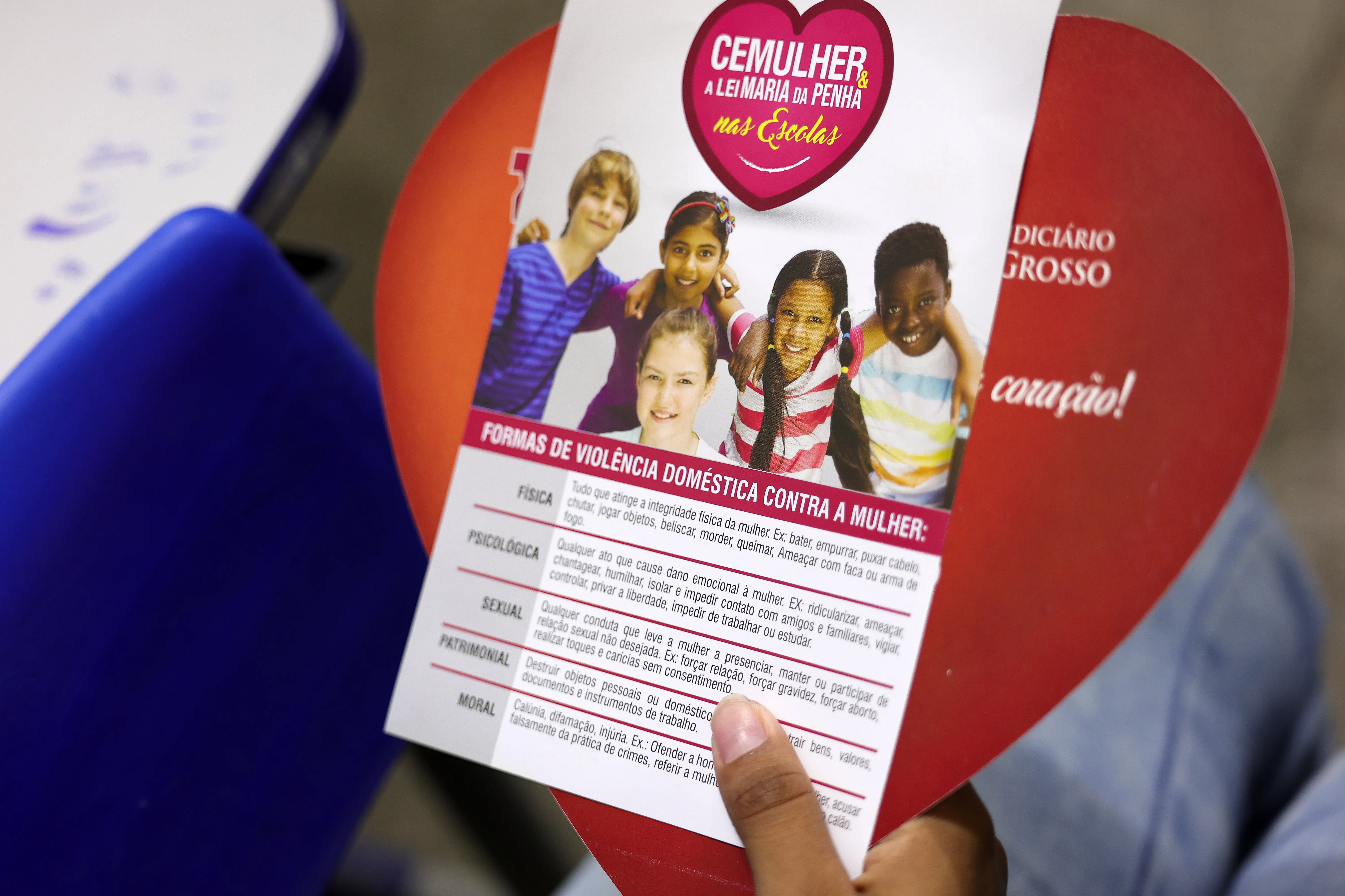 Mão segura folheto informativo com topo em formato de coração vermelho e textos sobre a Lei Maria da Penha e formas de violência doméstica, como física, psicológica, sexual, patrimonial e moral.