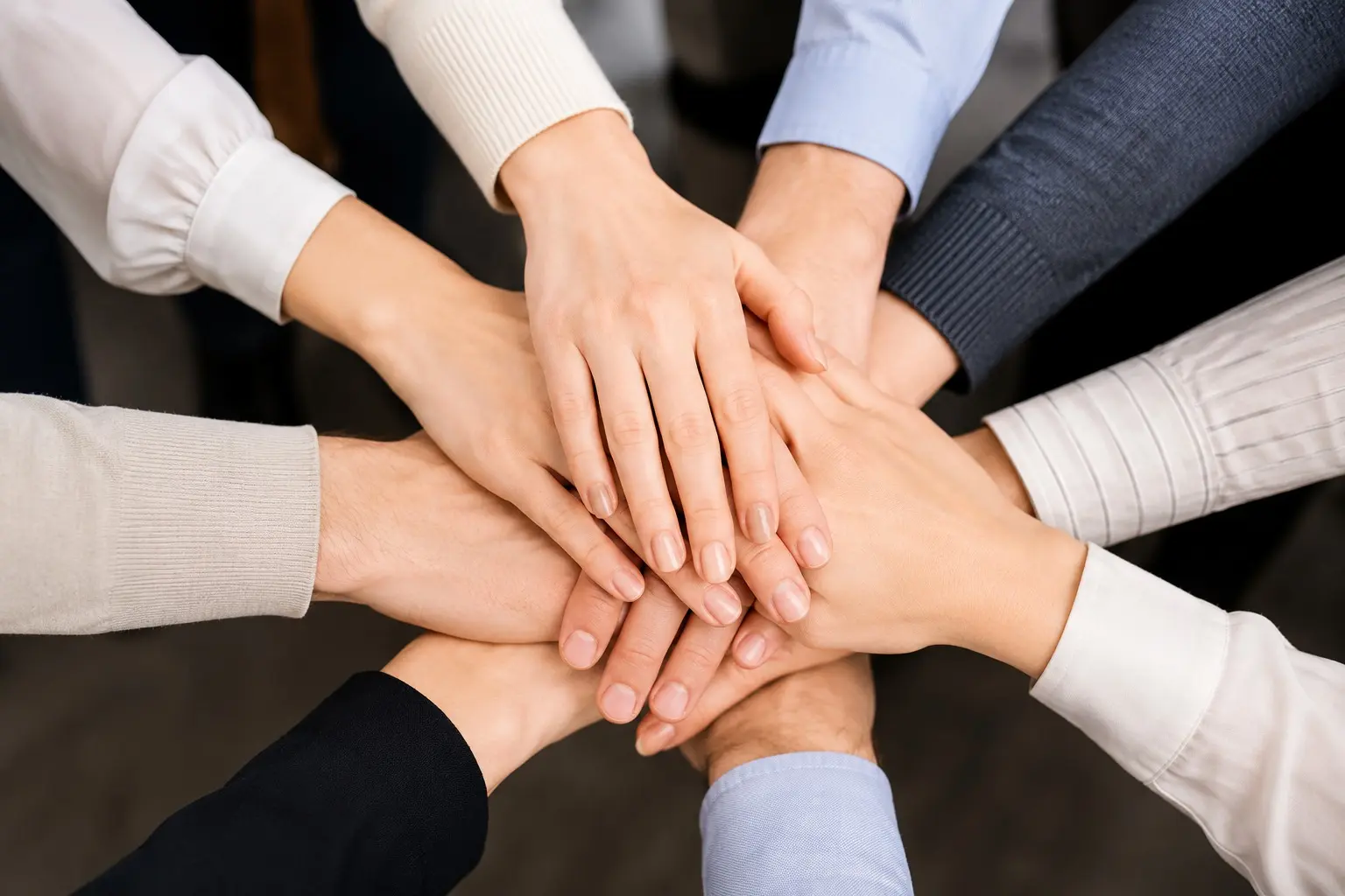 Mãos de pele clara (brancas) de várias pessoas vestindo camisas de manga longa estão sobrepostas e unidas no centro da imagem, formando uma pilha que simboliza trabalho em equipe.