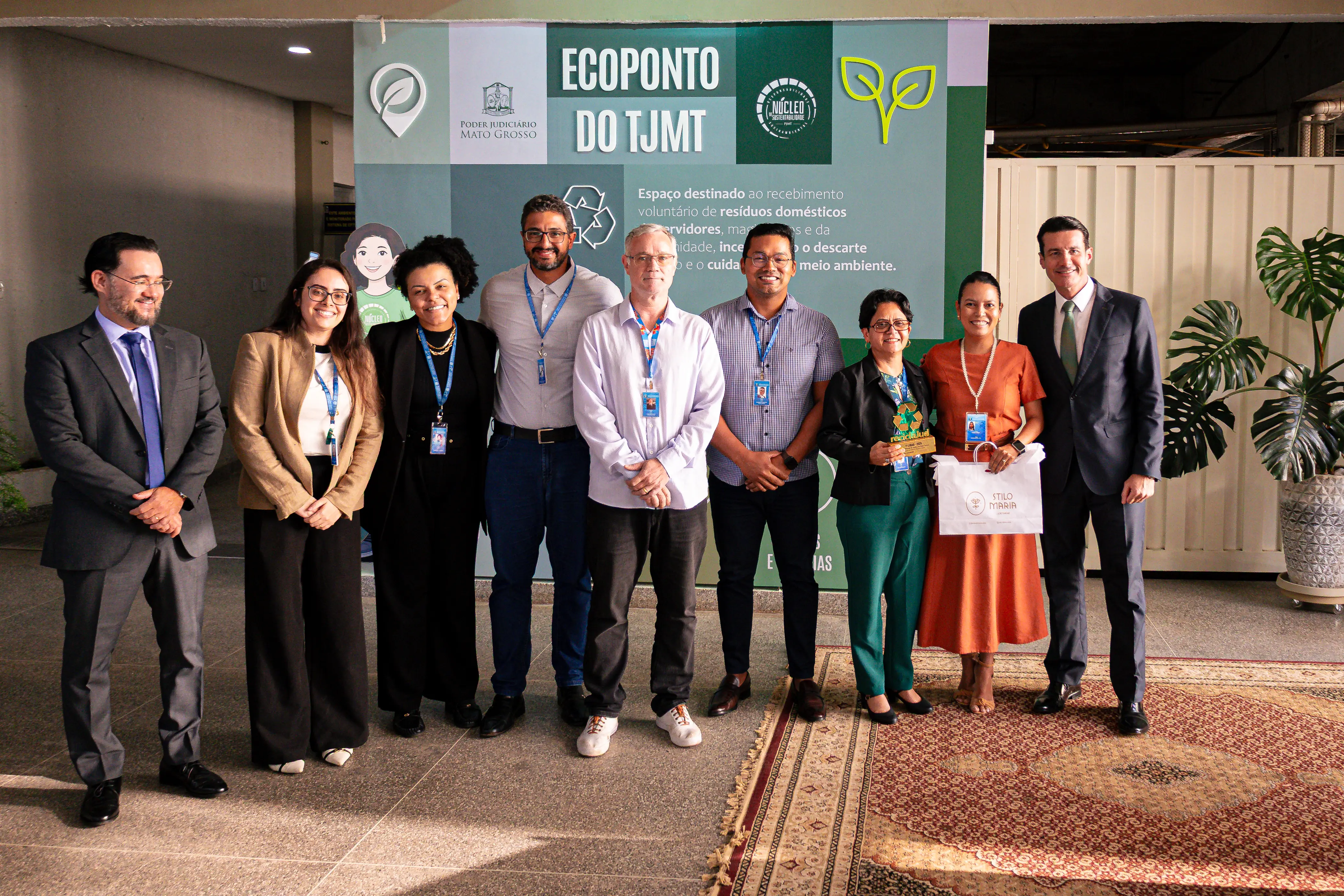 Uma nova fotografia posada em frente ao mesmo painel, agora com um grupo menor, composto por 9 pessoas (cinco homens e quatro mulheres). A formação é lado a lado e todos olham para a foto sorrindo. A maioria usa crachás no pescoço.