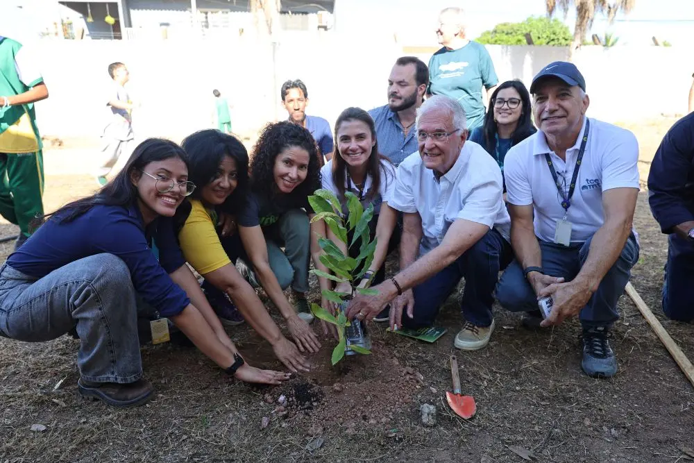 Foto horizontal que mostra quatro mulheres e três homens agachados ao solo, com as mãos estendidas para a muda de caju que eles plantaram no pátio da escola municipal Dom Bosco, em Cuiabá. Todos sorriem para a foto.