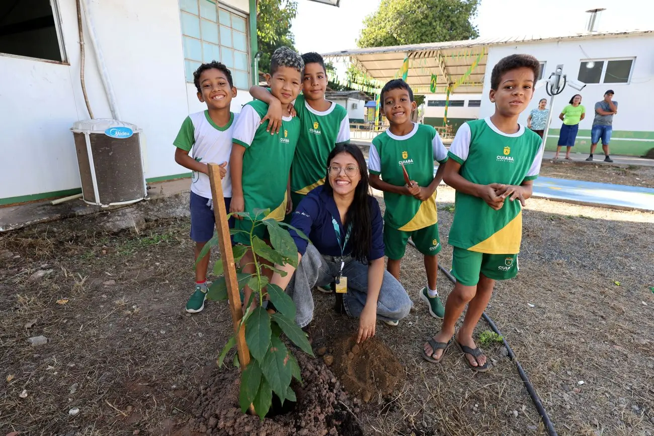 Foto horizontal em plano aberto que mostra cinco meninos de cerca de 8 anos de idade em pé, abraçados, e uma mulher com uniforme da Águas Cuiabá agachada ao solo, em frente a uma muda de árvores que eles plantaram no pátio da Escola Municipal Dom Bosco, no bairro Praeirinho, Cuiabá. Todos sorriem.