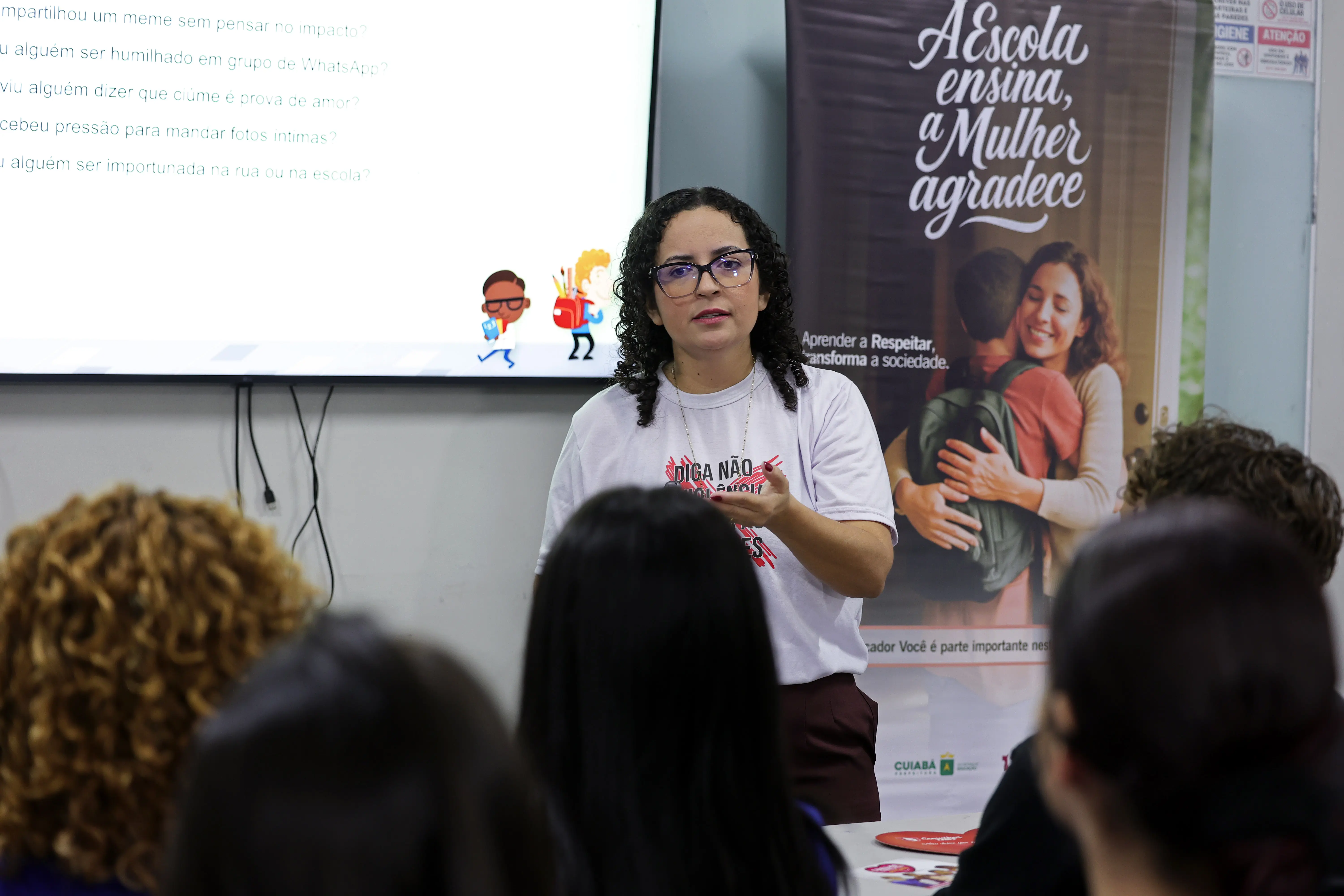 Mulher de óculos, cabelos cacheados e camiseta branca gesticula ao falar. Ao fundo, vê-se uma tela com perguntas e um banner com o texto 
