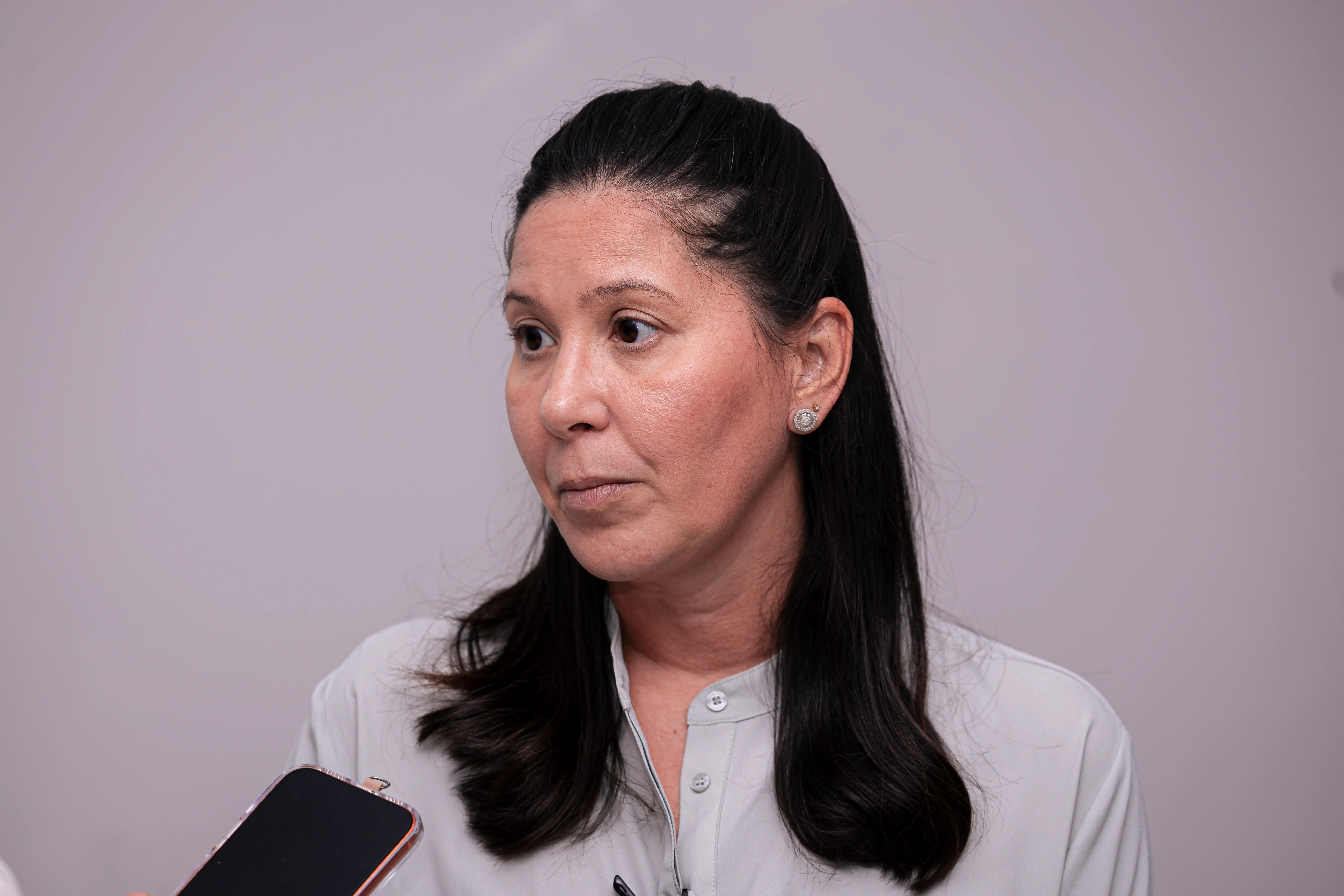 Mulher de pele clara e cabelos pretos presos em meio-preso, vestindo camisa cinza-clara, olha para a esquerda. No canto inferior esquerdo, aparece parte de um celular preto. Fundo cinza neutro.