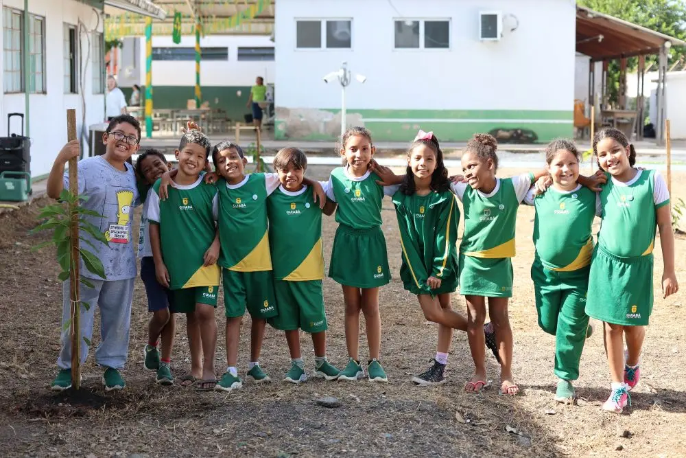 Foto horizontal em plano aberto que mostra um grupo de 10 alunos da EMEB Dom Bosco abraçados, em frene a uma muda de árvores plantada no pátio da escola. Os meninos e meninas têm cerca de oito anos de idade e todos usam uniforme verde e branco, exceto um menino com calça e camiseta cinza.