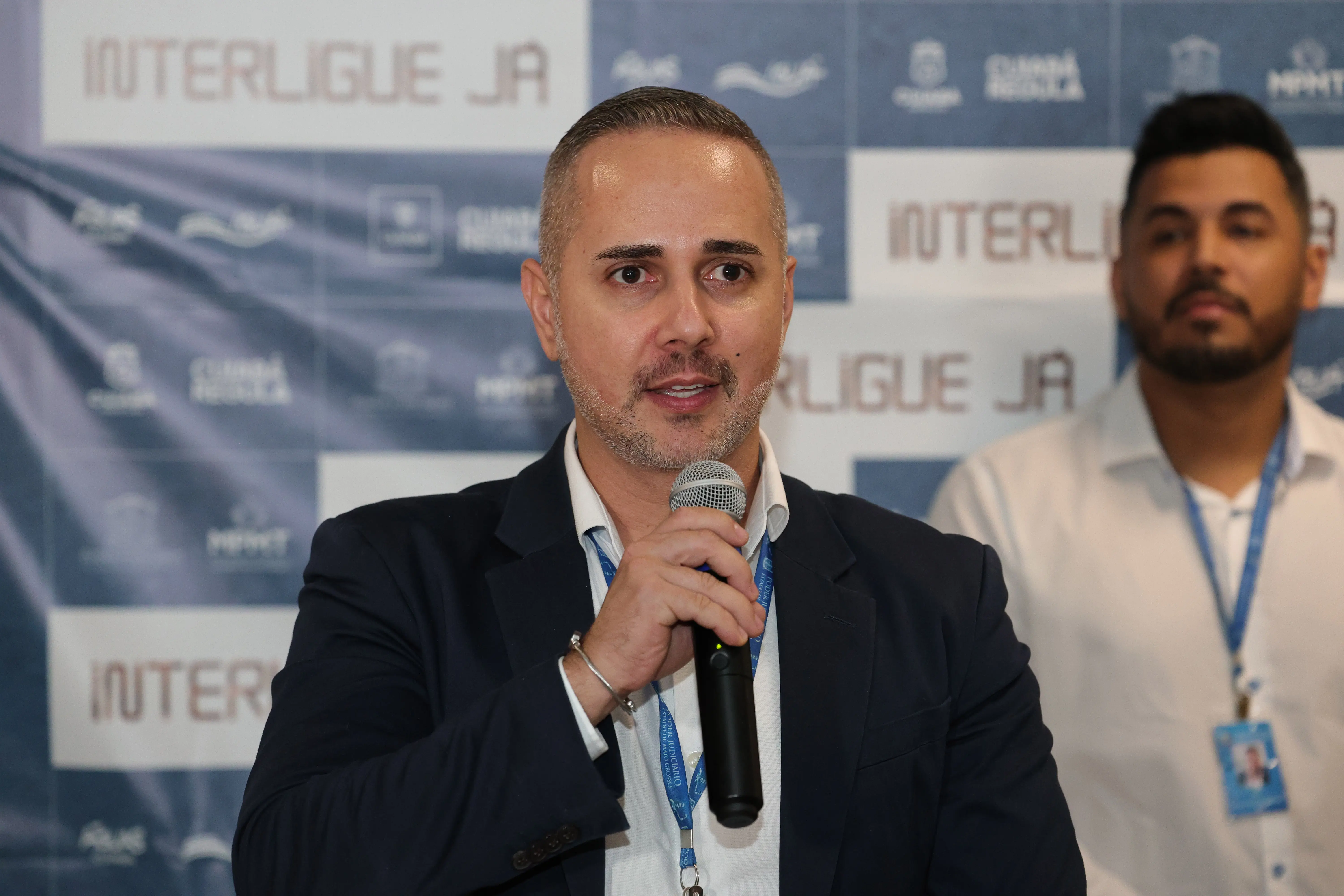 Homem de cabelos grisalhos curtos, terno azul-marinho, fala ao microfone seguro com a mão. Ao fundo, painel do 