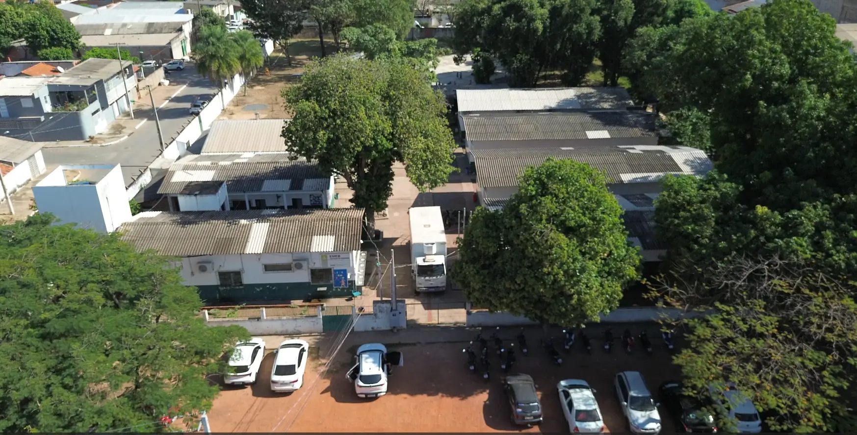 Foto horizontal que mostra vista aérea da Escola Municipal Dom Bosco, no bairro Praeirinho, em Cuiabá. O prédio da unidade é rodeado de árvores altas e há carros estacionados na frente.