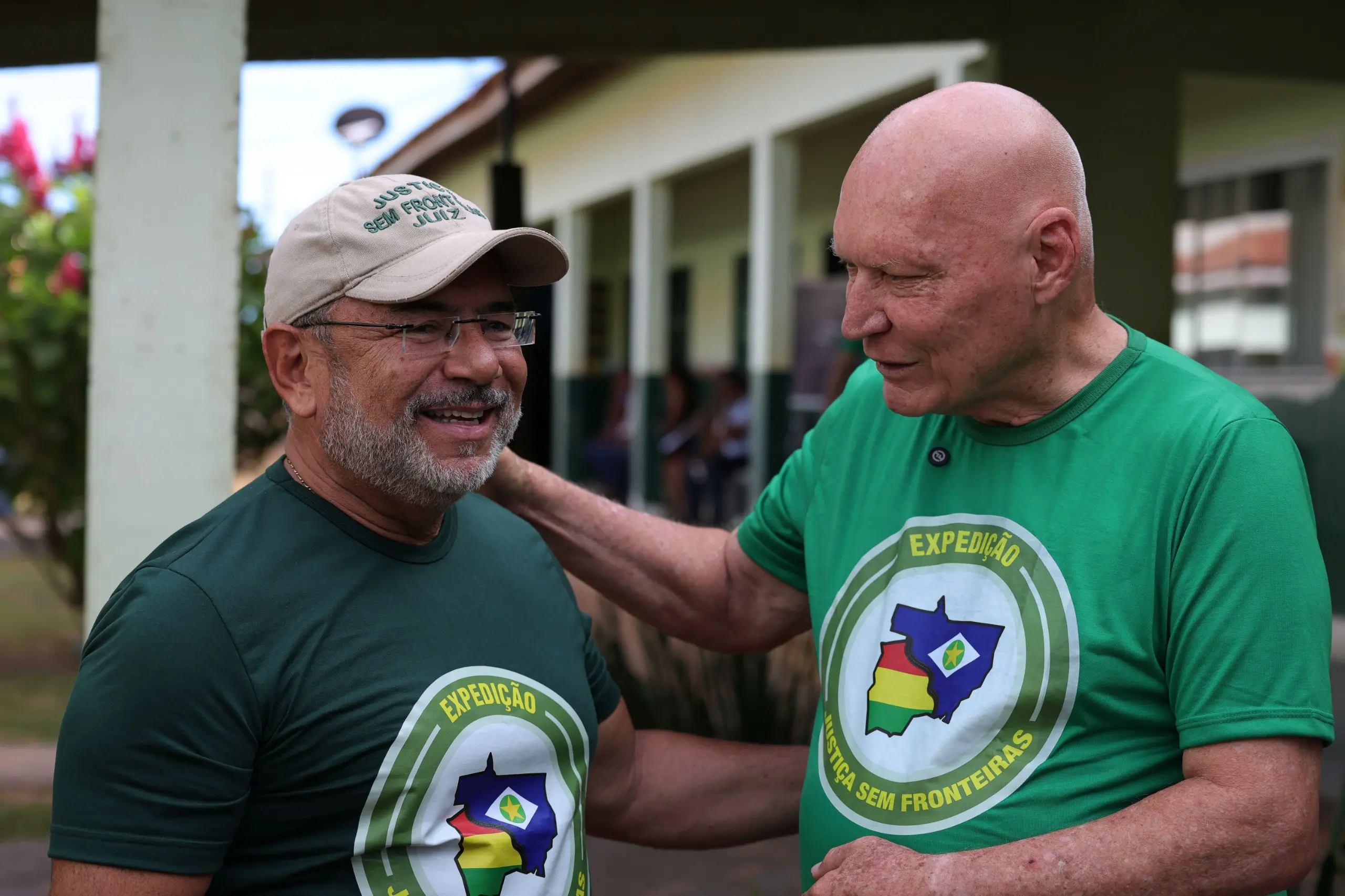 O homem de camiseta verde-clara abraça amigavelmente pelos ombros o homem de barba grisalha, óculos e boné bege. Ambos sorriem em um ambiente externo com um prédio ao fundo.