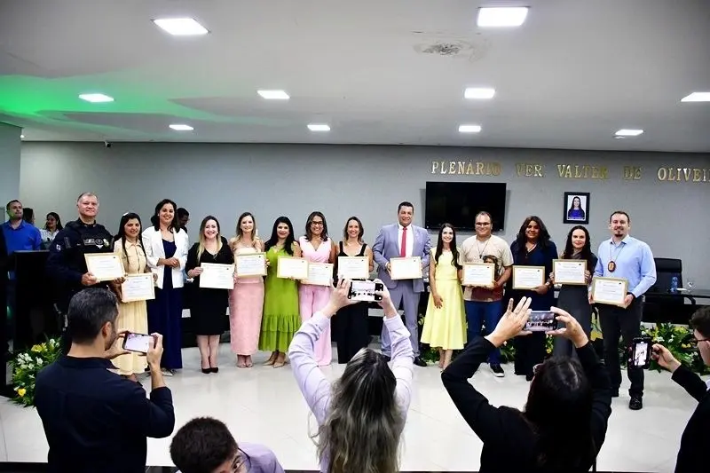 Amplo grupo de homens e mulheres em traje social posa em um plenário, segurando certificados. Em primeiro plano, pessoas fotografam o momento com celulares.