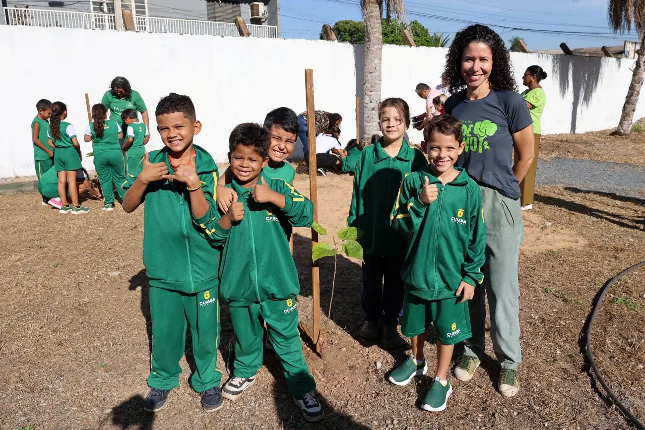 Foto horizontal em plano aberto que mostra quatro meninos e uma menina com uniforme verde da escola municipal Dom Bosco e a engenheira florestal Rosiani Carnaíba, com camiseta do programa Verde Novo, sorrindo para a foto, em pé, em frente a muda de árvore que plantaram no pátio da escola.