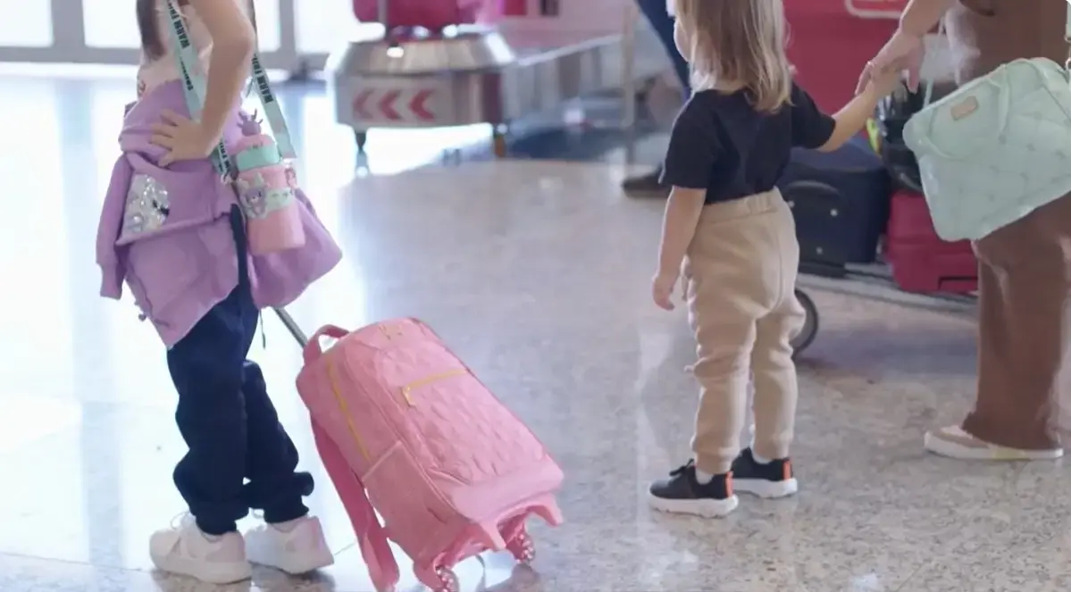Foto horizontal que mostra duas meninas de costas, em um aeroporto. A maior, com cerca de 8 anos, aparece do busto para baixo, segurando uma mochila de rodinhas cor-de-rosa. A menor, com cerca de 2 anos, está segurando a mão de uma mulher.