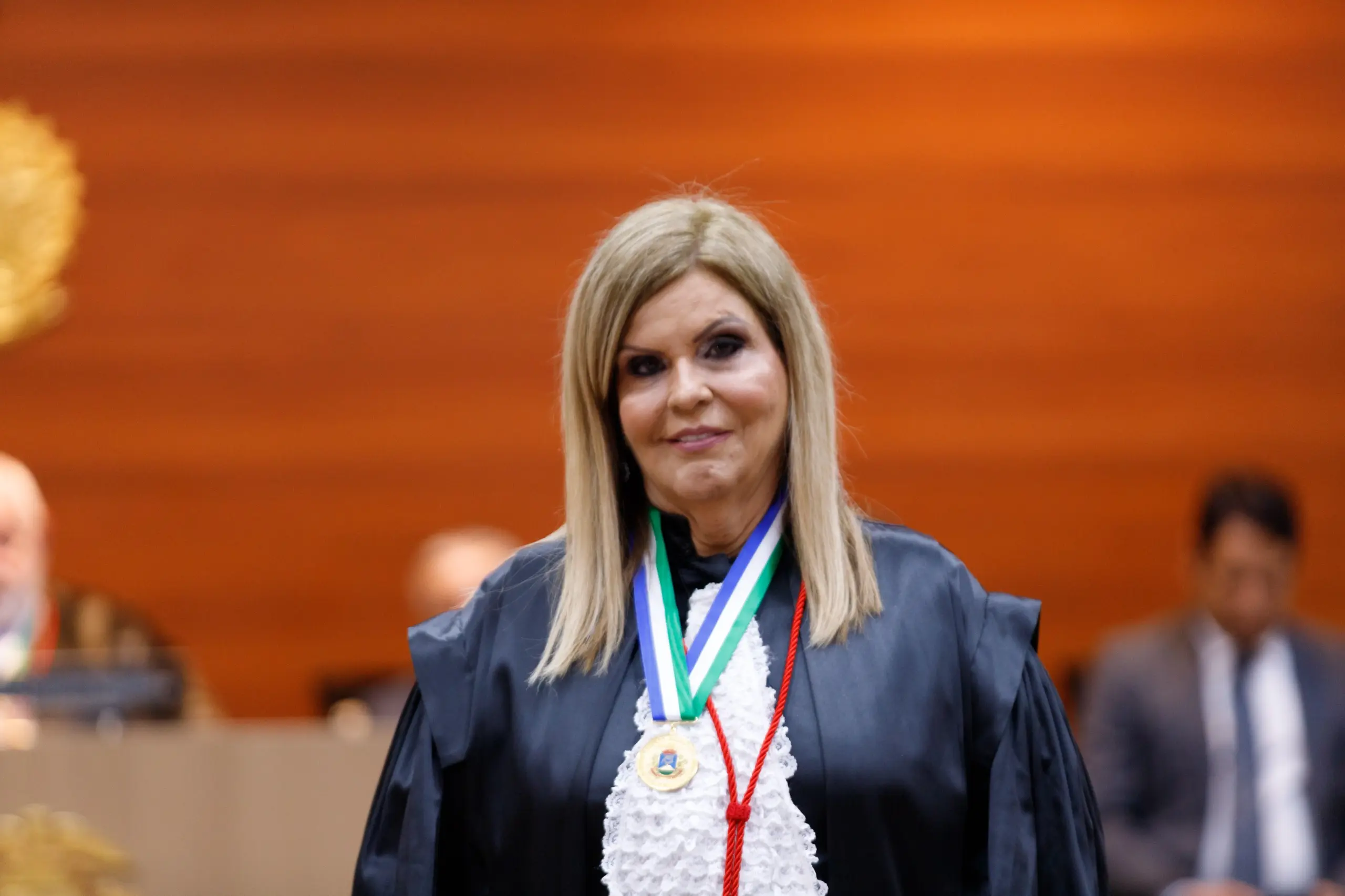 Foto horizontal colorida da vice-presidente Nilza Pôssas de Carvalho. El é uma mulher loira, de cabelos lisos abaixo dos ombros. Usa toga de gala preta com babados brancos na frente e medalha envolta do pescoço