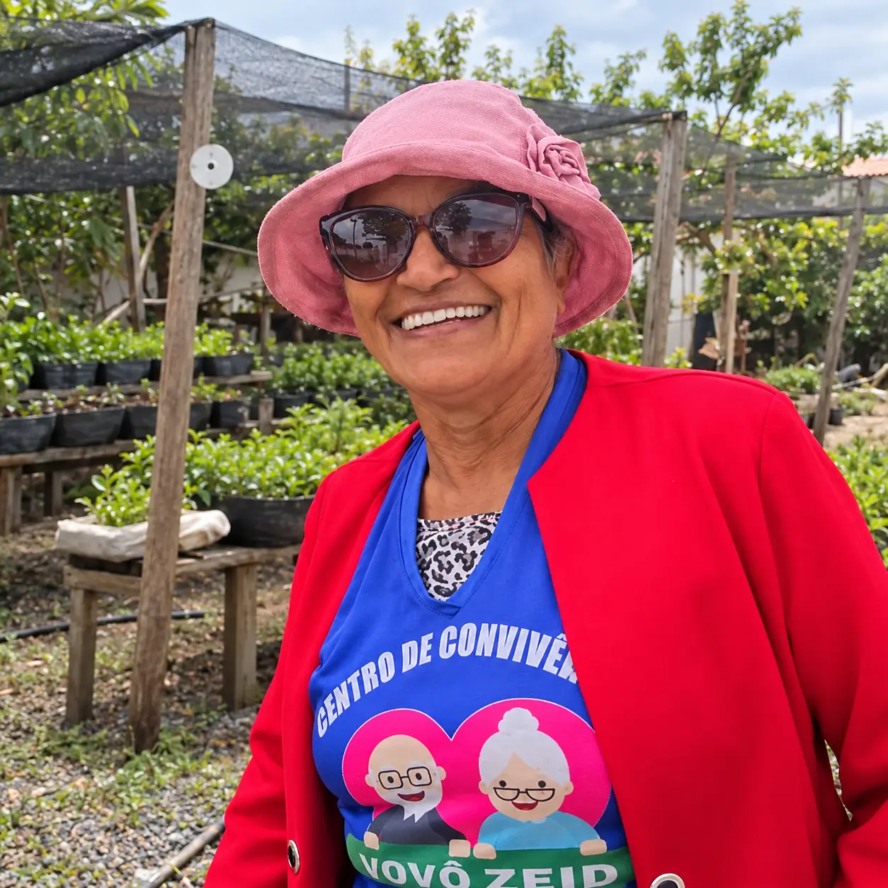 Foto colorida de uma mulher idosa sorrindo em uma horta, usando chapéu rosa, óculos escuros, casaco vermelho e avental azul do Centro de Convivência Vovô Zeid.