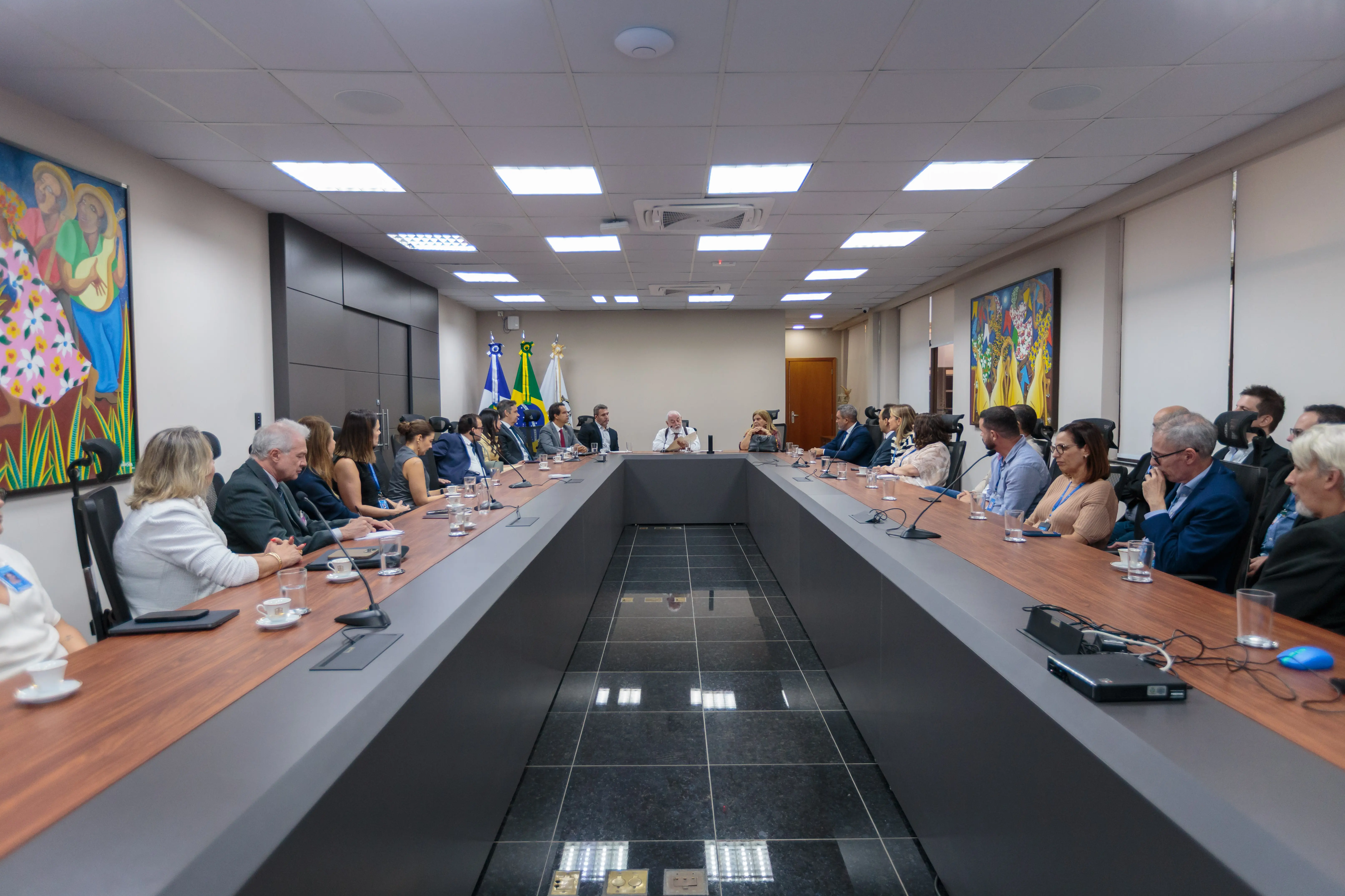 A imagem mostra toda a sala de reuniões, cerca de 30 pessoas sentadas em volta da mesa de reunião. Na cabeceira da mesa estão o presidente do TJMT, a vice-presidente e o desembargador Luiz Saboia