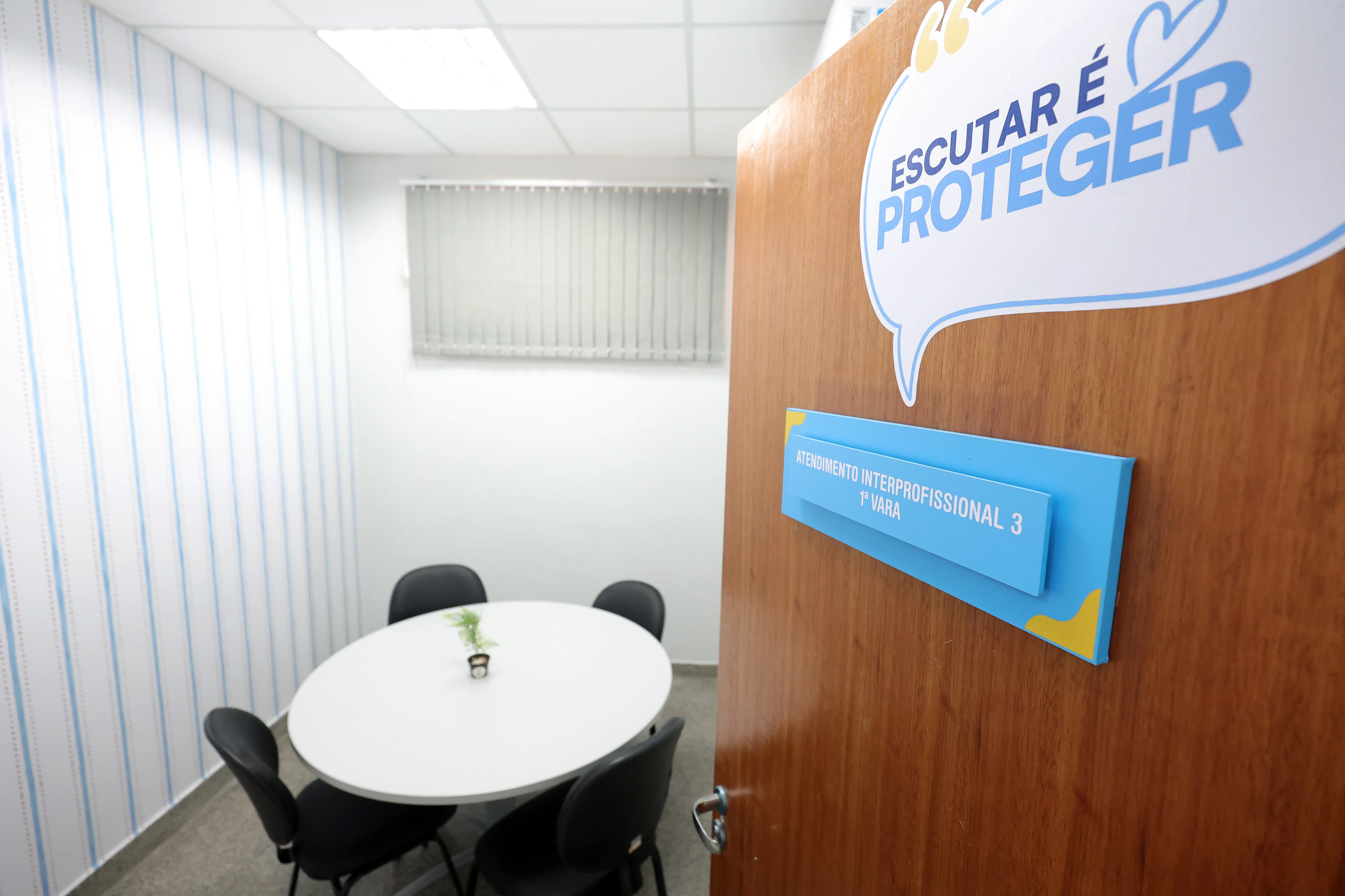 Visão parcial de uma porta de madeira aberta com a placa "Atendimento Interprofissional 3". Acima um balão com a frase "escutar é proteger". Ao fundo, vê-se uma pequena sala com mesa redonda branca e quatro cadeiras pretas.