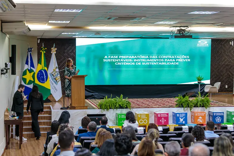 Mulher discursa em púlpito num palco de auditório ao lado de um grande painel verde. À sua frente, no chão, há cubos coloridos dos ODS e uma plateia atenta que assiste ao evento.