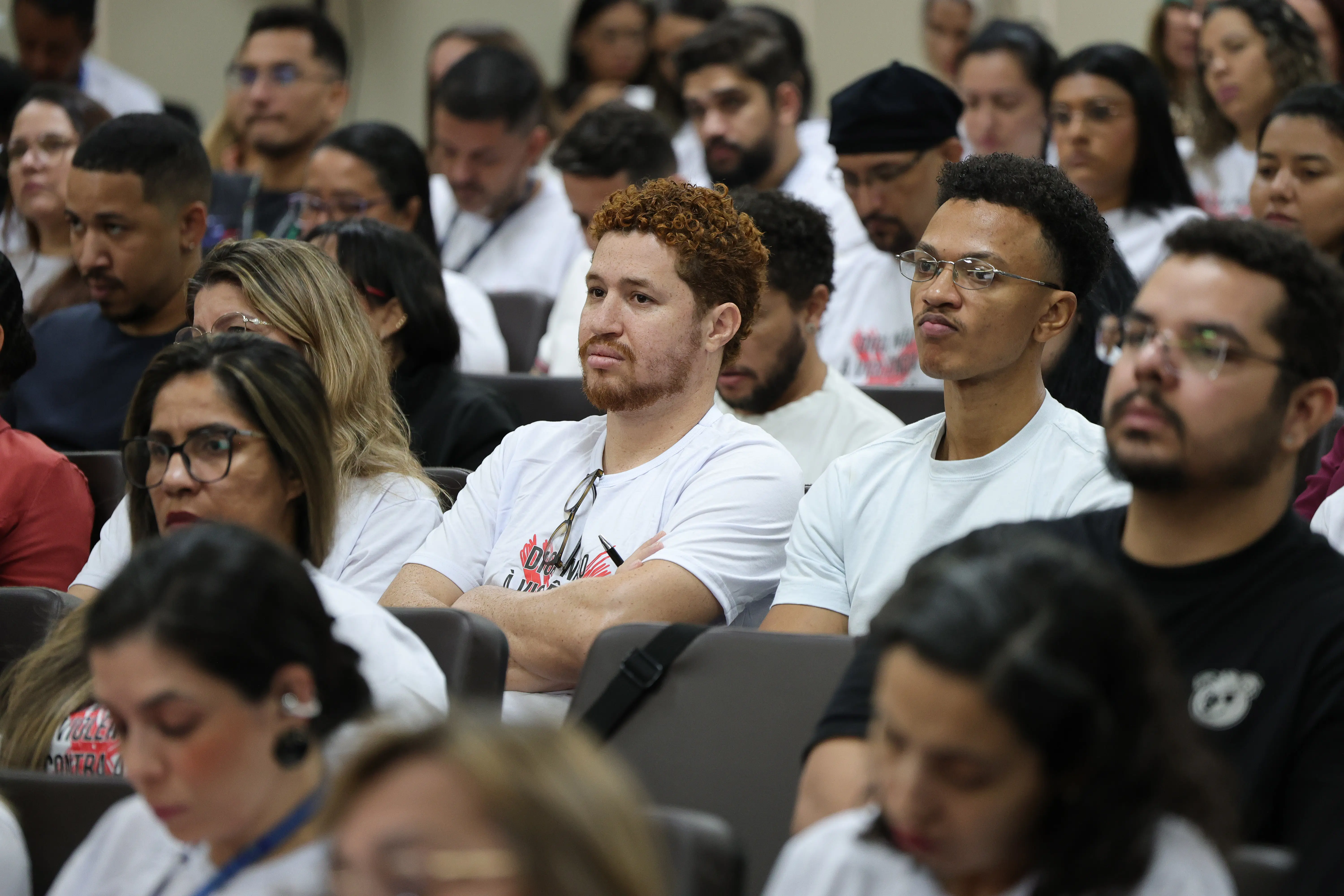 Plano médio da plateia focando em dois homens jovens sentados lado a lado: um de cabelo cacheado ruivo e braços cruzados, e outro de óculos e cabelo curto escuro. Ambos vestem camisetas brancas e olham para a frente.
