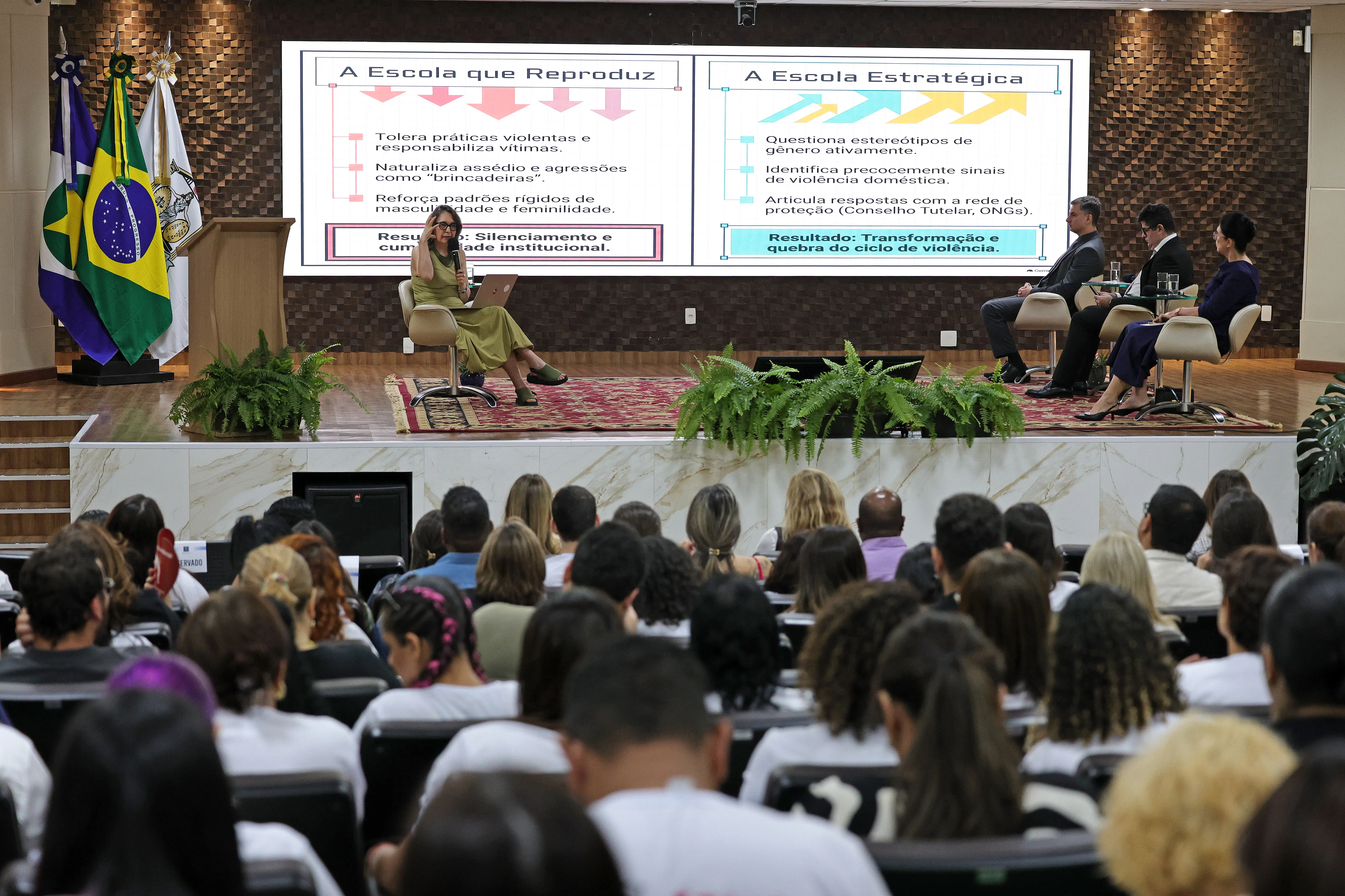 Visão geral do auditório lotado, de costas para o público, em direção ao palco. No palco, uma palestrante sentada com notebook apresenta slides em uma grande tela ao fundo sobre "A Escola que Reproduz" e "A Escola Estratégica". Outras três pessoas estão sentadas no palco.