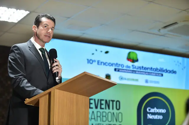Homem de terno escuro e gravata verde fala ao microfone em púlpito de madeira. Ao fundo, painel com o texto "10º Encontro de Sustentabilidade" e o selo "Carbono Neutro".