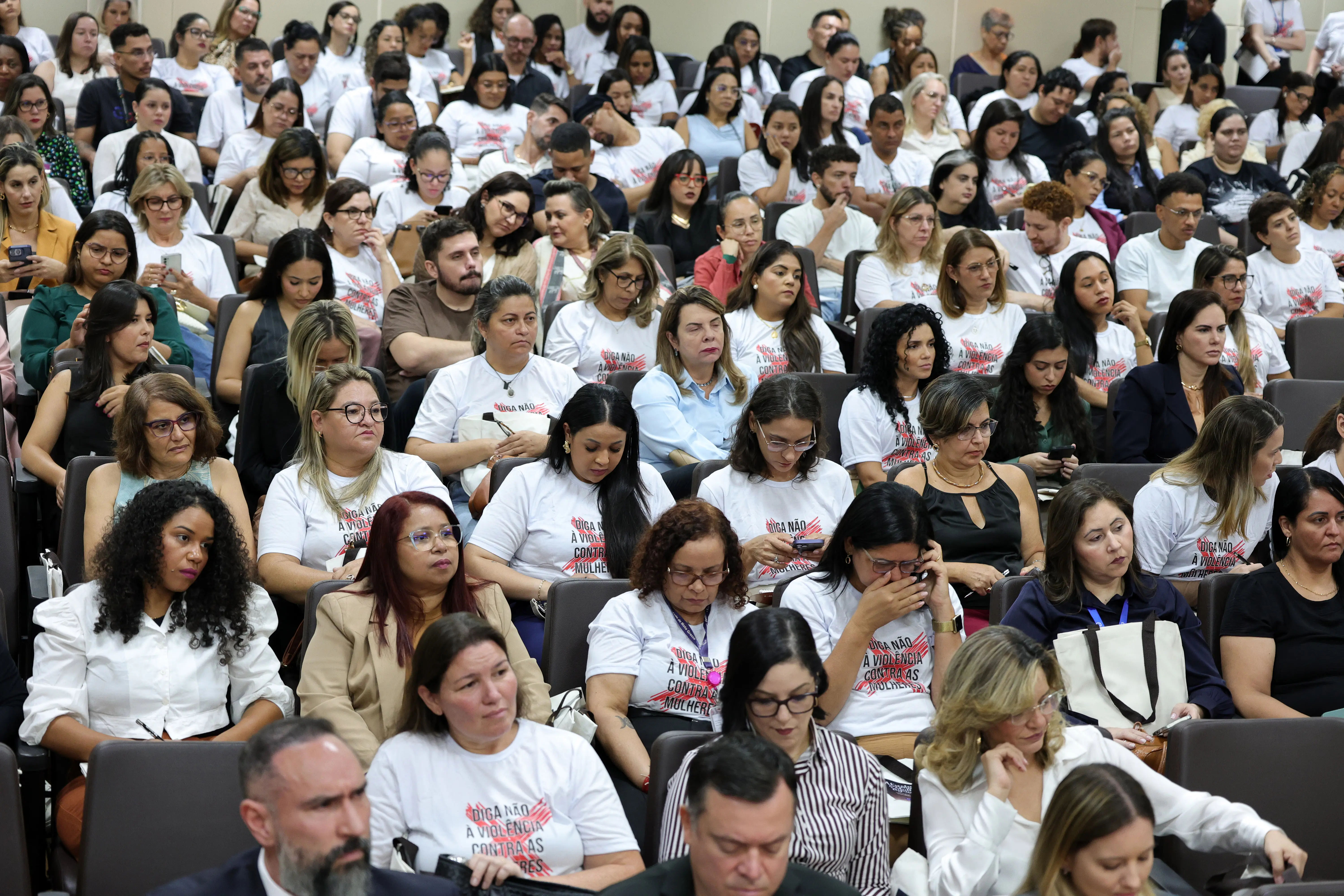 Plenária cheia de pessoas sentadas em um auditório. Várias delas vestem camisetas brancas com a frase "Diga não à violência contra as mulheres". A maioria das pessoas observa atentamente em direção ao palco.