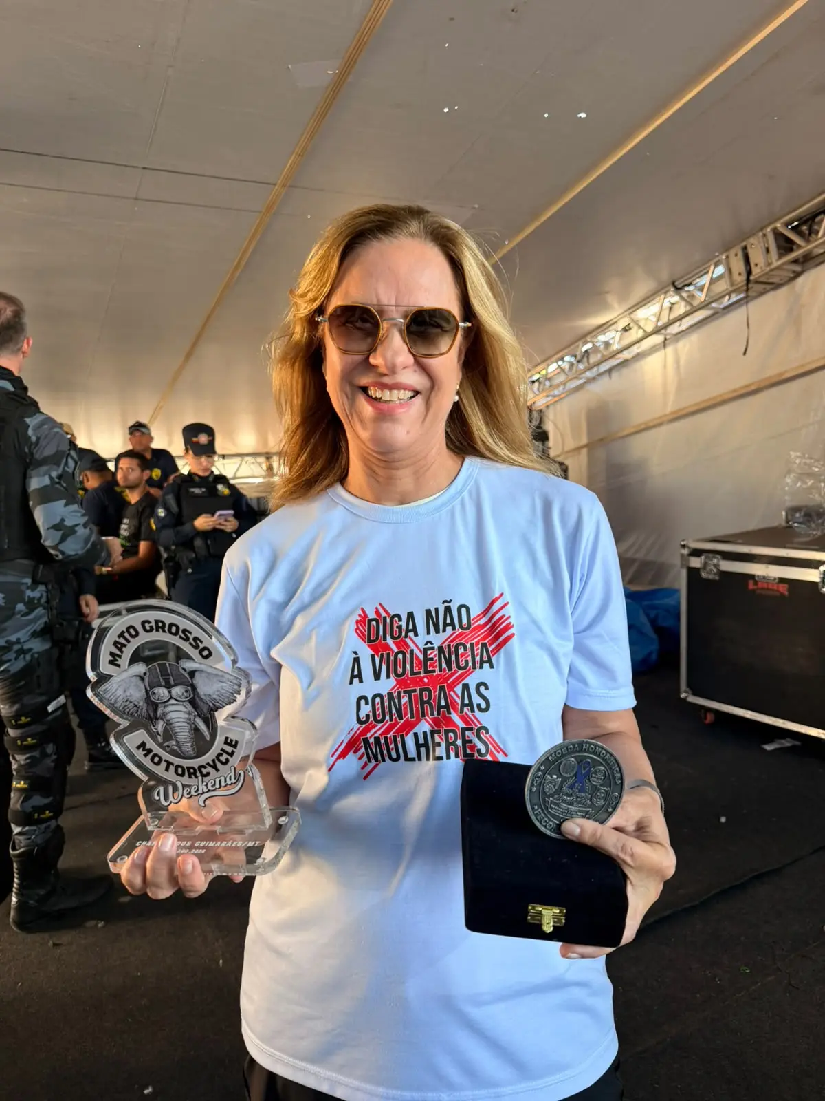 Foto da desembargadora Vandymara sorrindo para a foto e segurando troféu e moeda honorífica referente ao Encontro de Motociclistas. Ela usa camiseta branca com estampa que diz "Diga não à violência contra as mulheres".