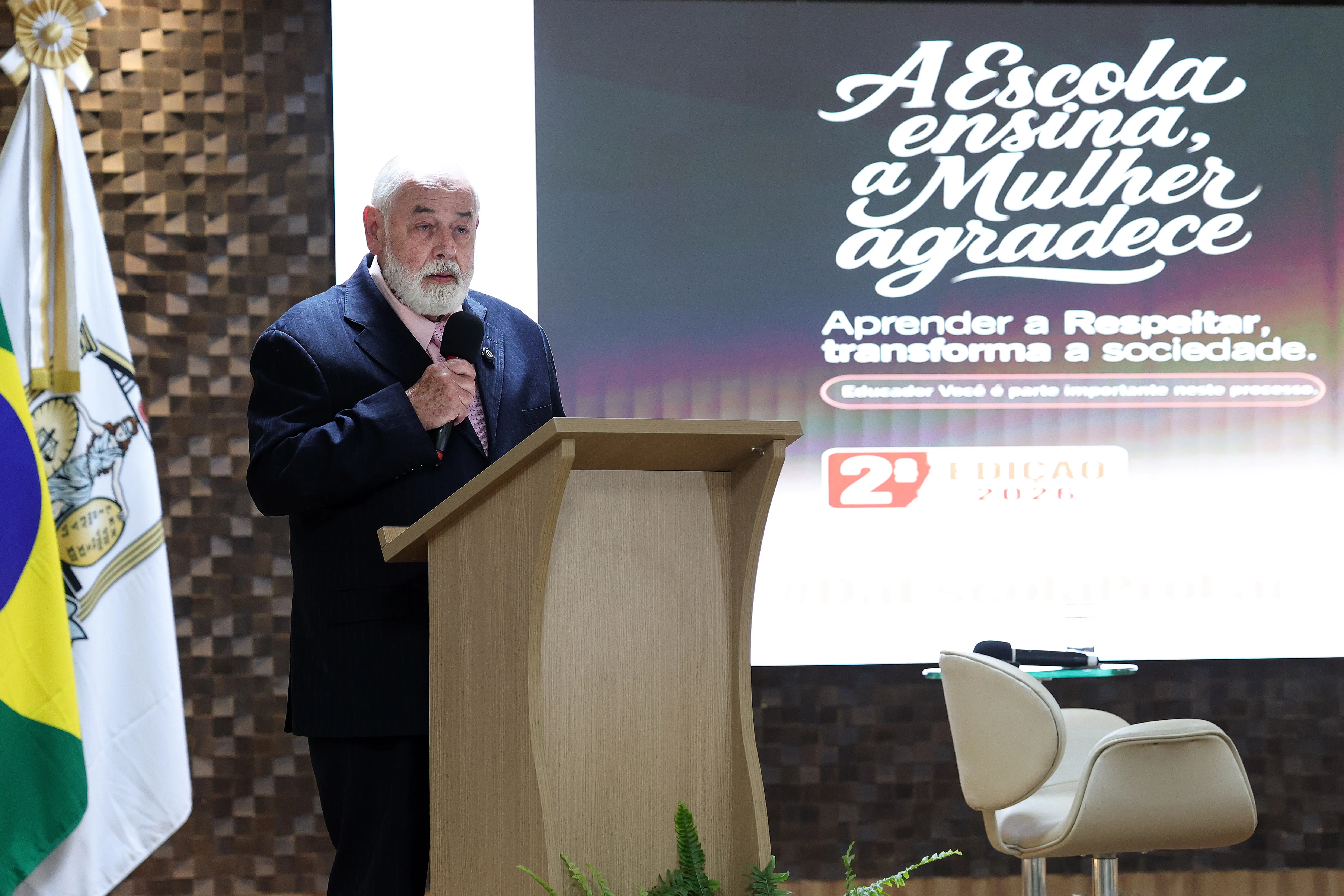Homem idoso, de barba branca e terno escuro, fala ao microfone em um pódio de madeira. À esquerda, uma bandeira do Brasil. Ao fundo, uma tela exibe a frase "A Escola ensina, a Mulher agradece" e a mensagem "Aprender a Respeitar, transforma a sociedade. 2ª Edição 2026".