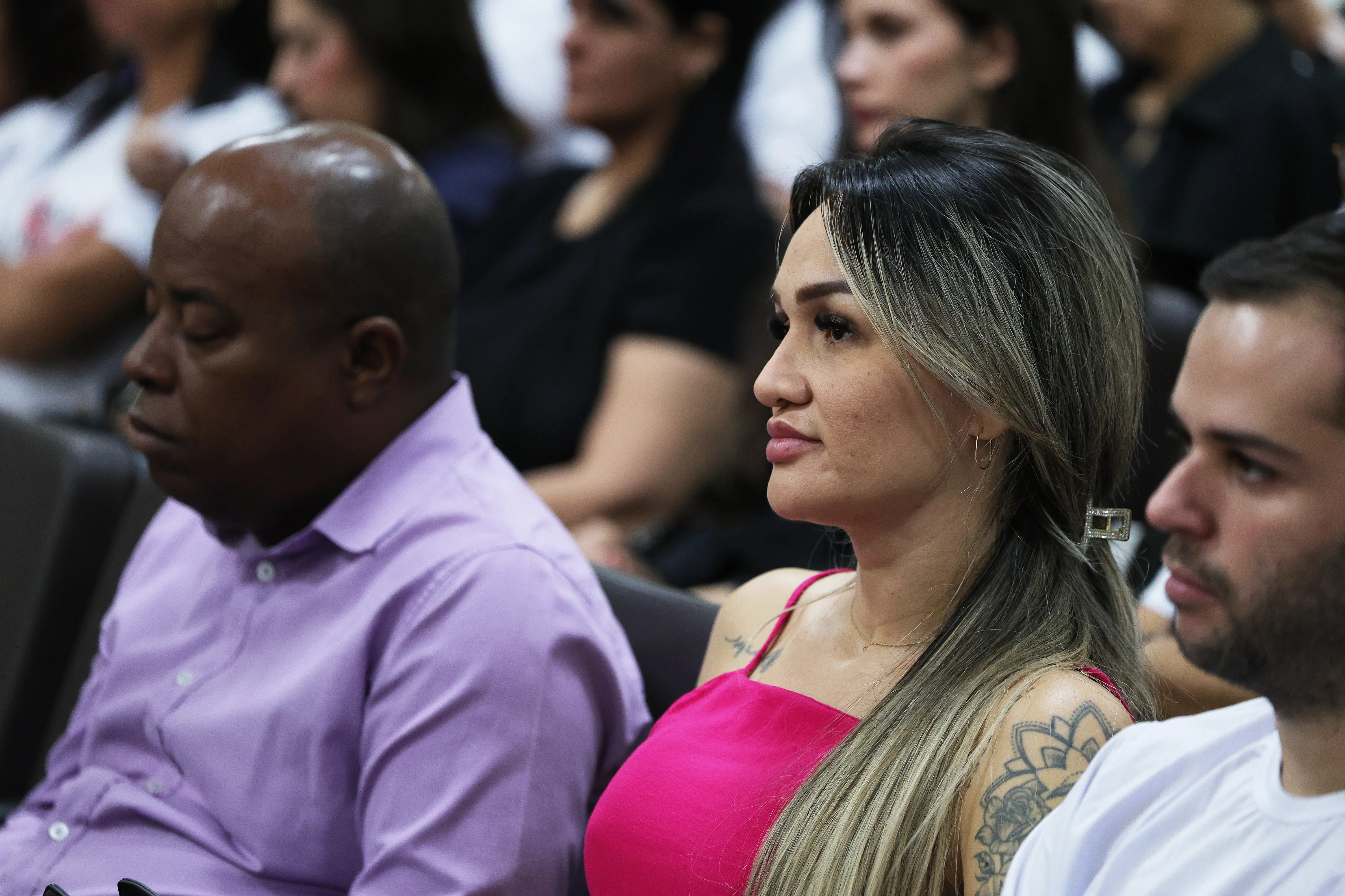 Plano fechado de perfil de uma mulher de cabelos loiros e compridos, vestindo blusa rosa de alça e com tatuagem no braço, sentada na plateia do auditório. Ao lado dela, um homem de camisa social lilás olha para a frente.