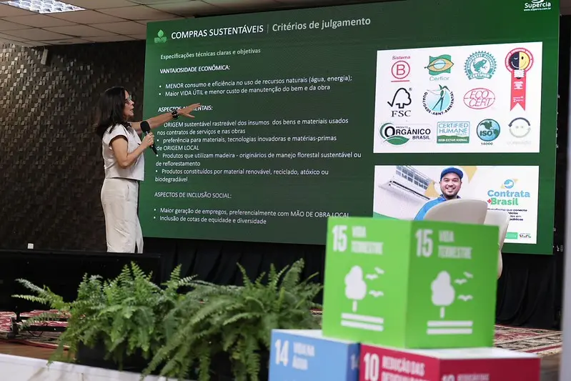 Mulher de roupa clara apresenta palestra ao lado de grande tela sobre "COMPRAS SUSTENTÁVEIS". Em primeiro plano, samambaias e cubos coloridos decorativos com o símbolo dos ODS.