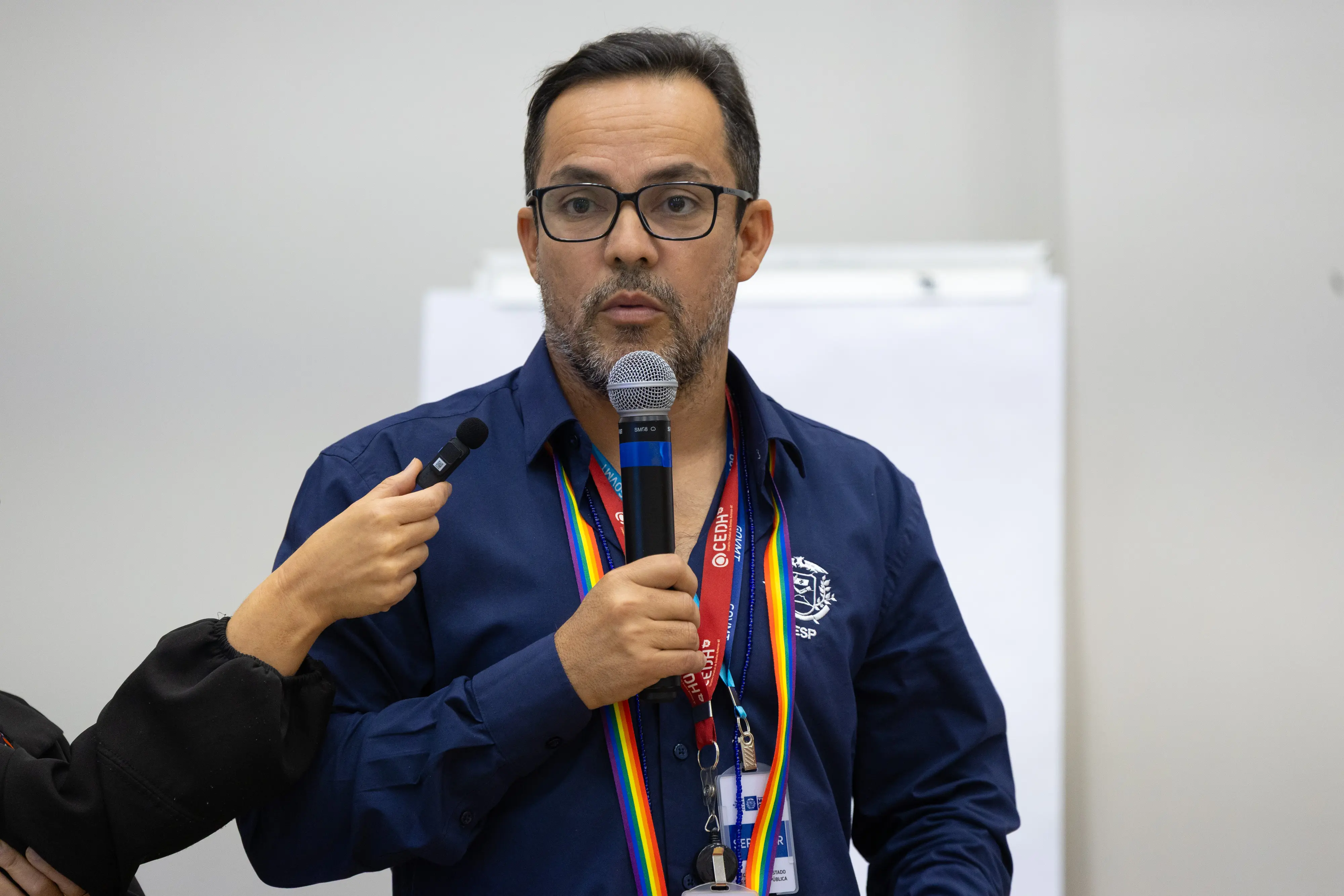 Homem de óculos fala ao microfone durante atividade da oficina. Participante segura gravador próximo ao entrevistado.