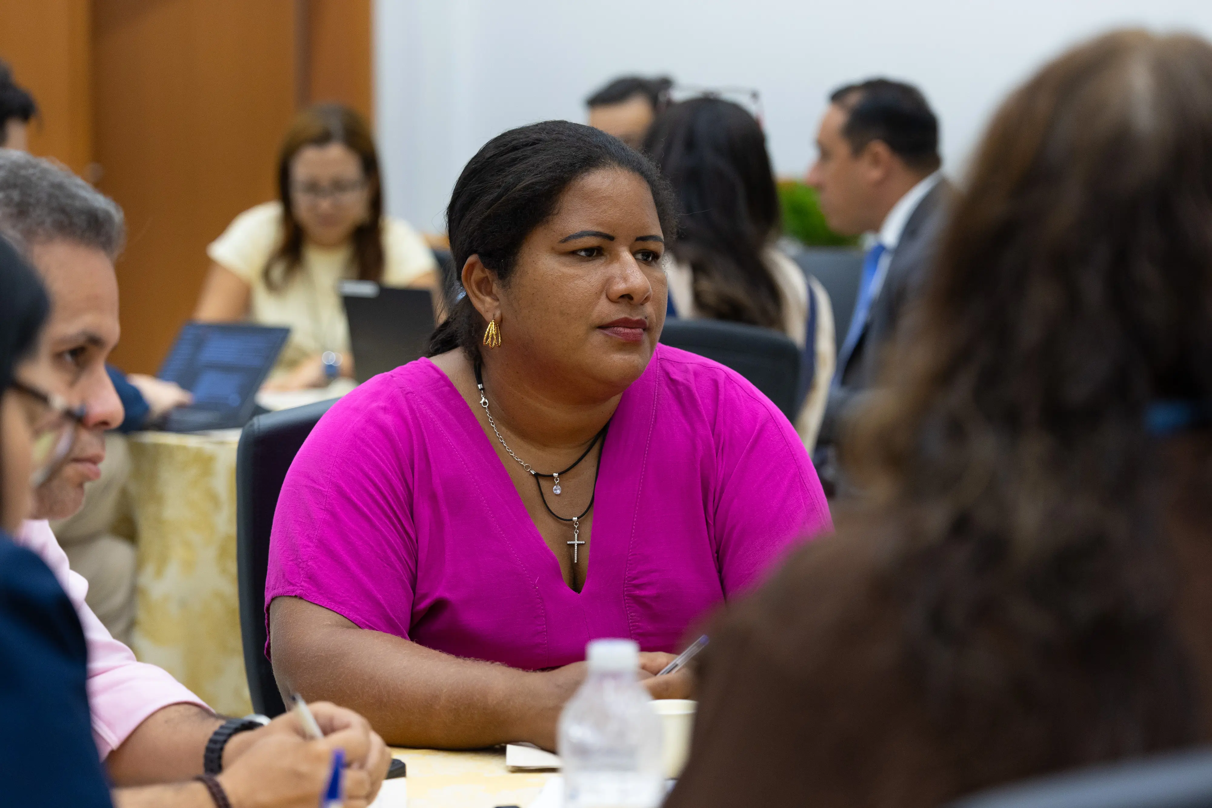 Mulher de blusa rosa participa de roda de conversa durante oficina. Pessoas aparecem desfocadas ao redor.
