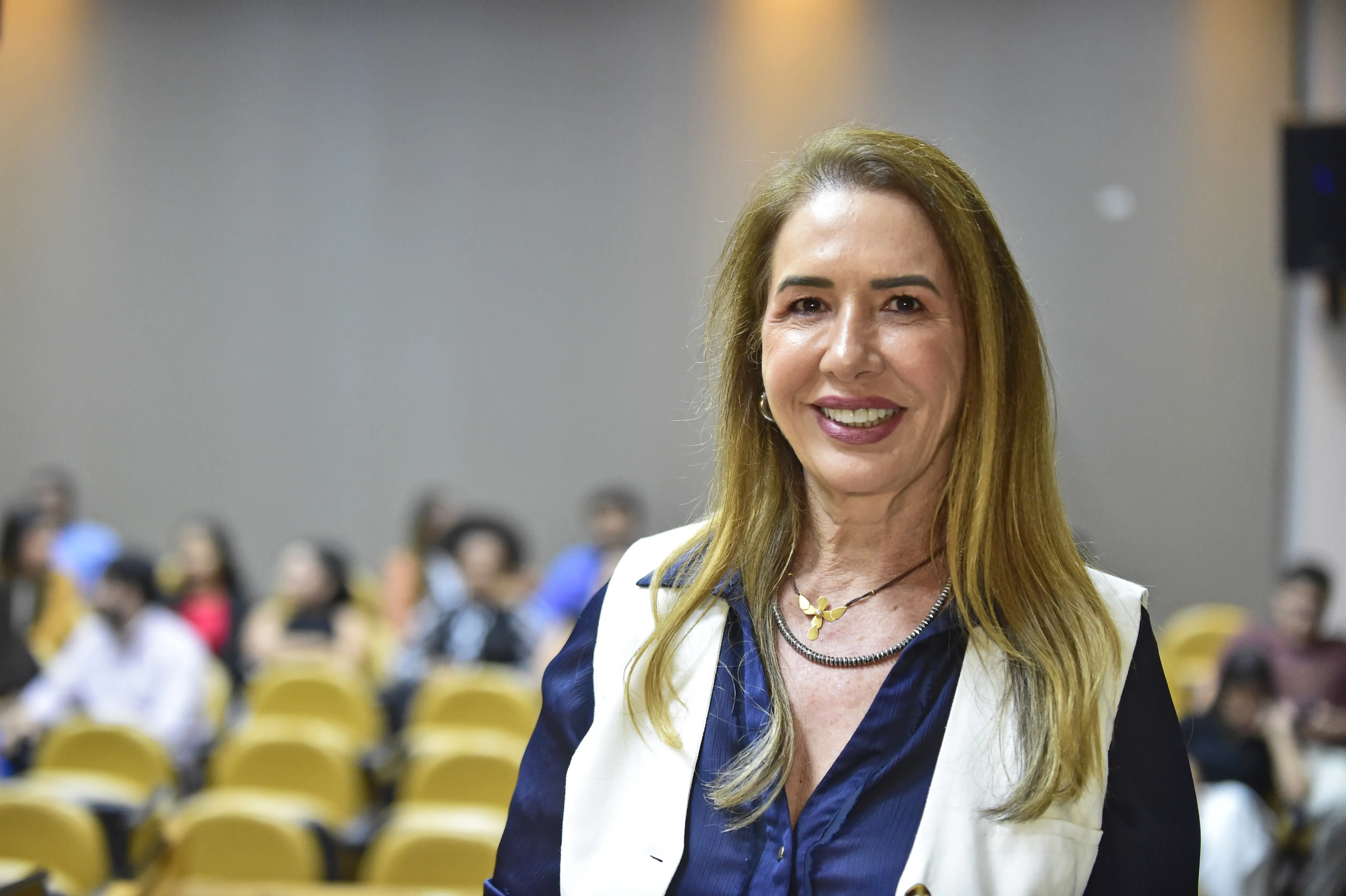 Uma mulher sorri, vestindo uma blusa azul com um colete branco por cima e um colar. Ela tem cabelo loiro e comprido. Ao fundo, cadeiras amarelas e pessoas desfocadas em um auditório. 